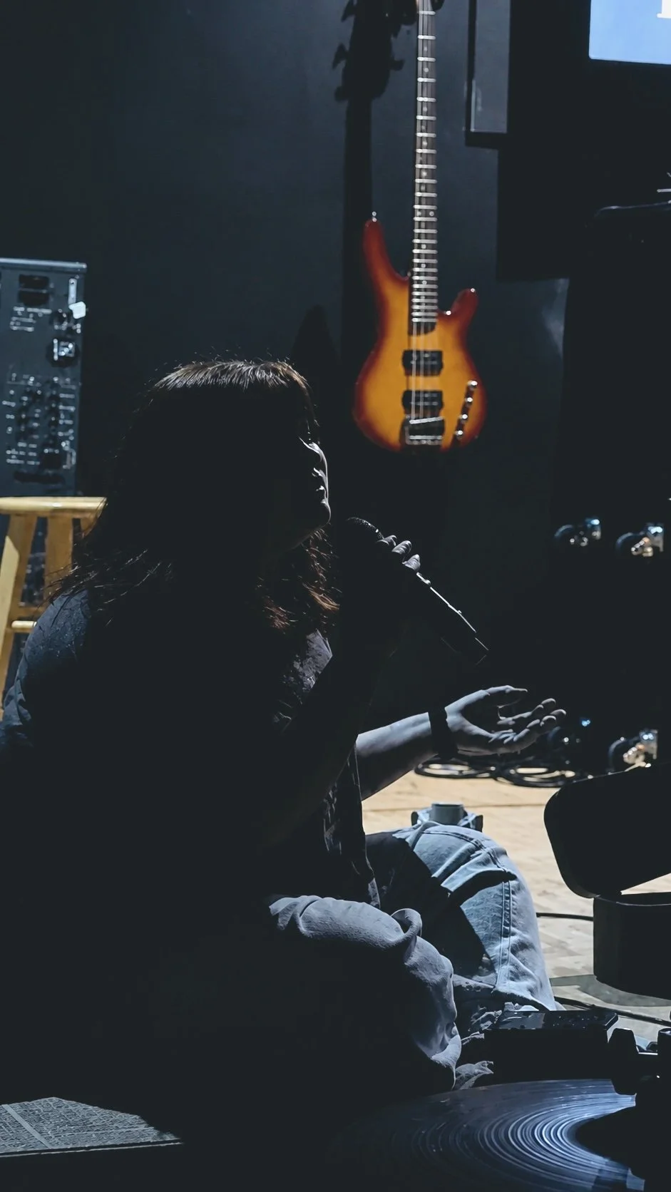 A woman with long hair sitting on the floor, speaking or singing into a microphone, with musical equipment and an electric guitar hanging on a dark wall in the background.