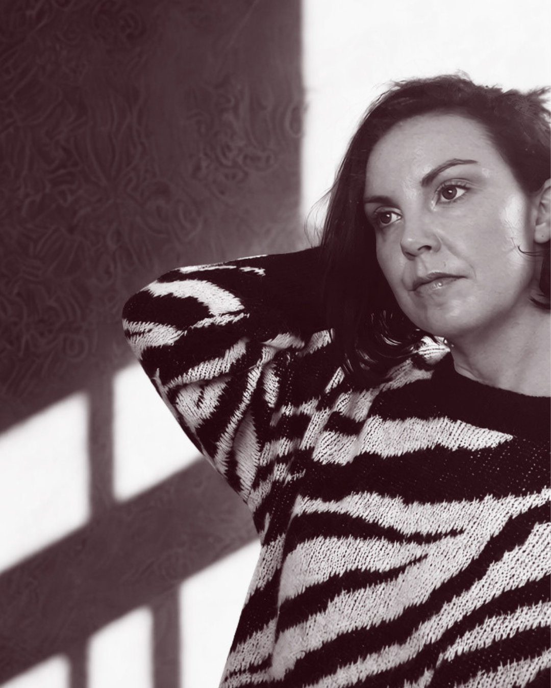 Black and white photo of a woman with dark hair, wearing a zebra print sweater, resting her head on her hand, with shadows cast on the wall behind her.