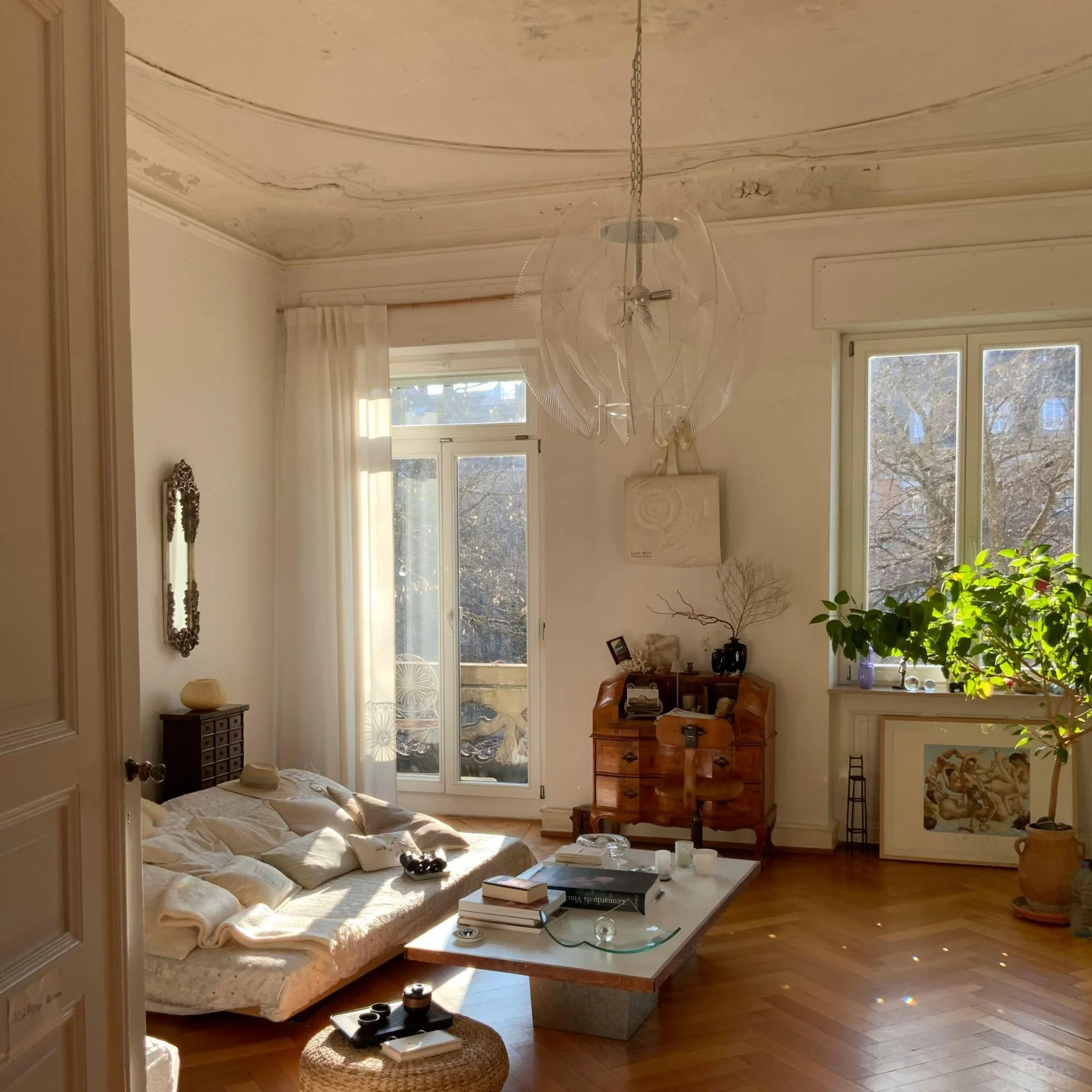 Sunlight streaming into a cozy living room with high ceilings, large windows, a beige couch with pillows, a wooden coffee table with books and candles, a tall green plant, and artwork on the walls.
