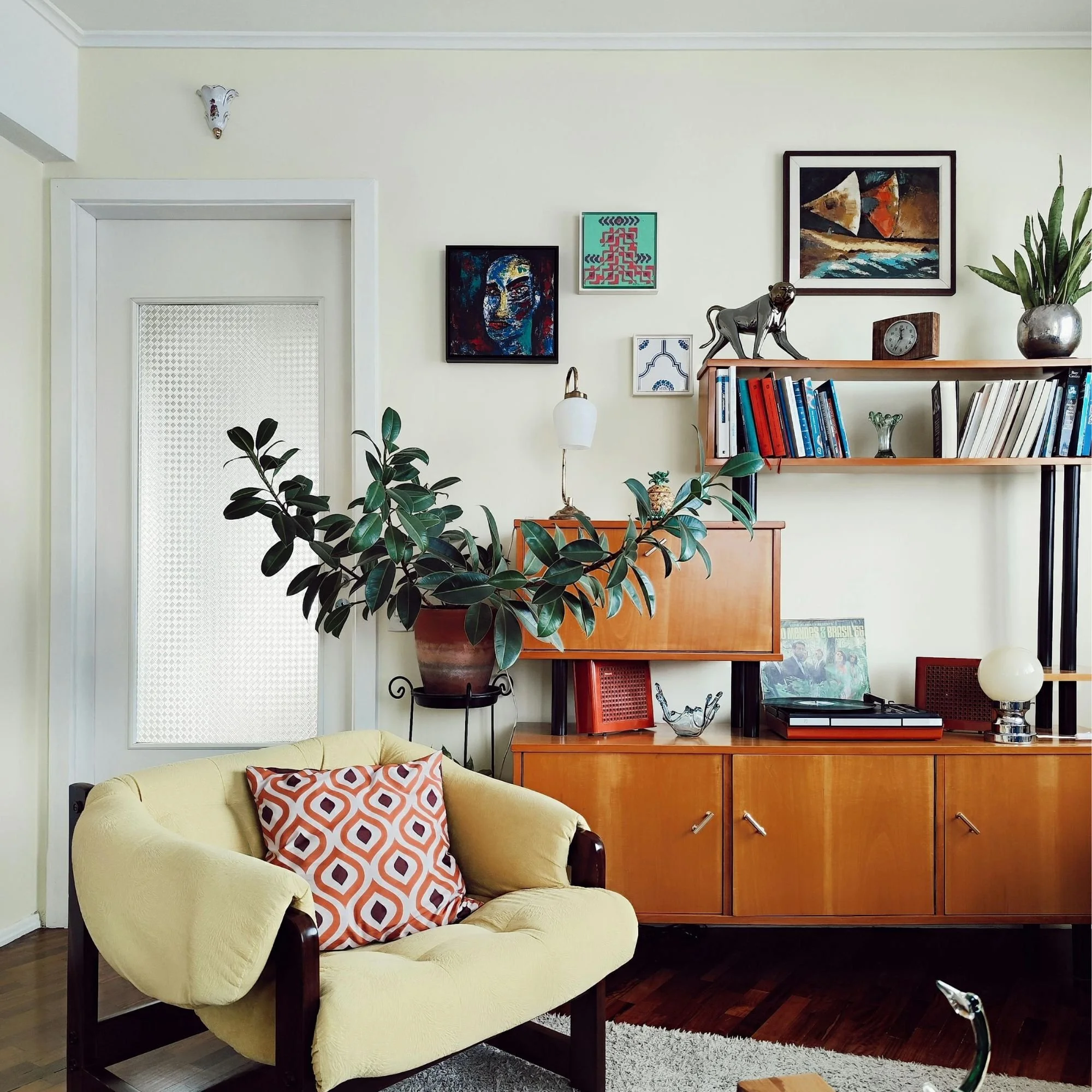 Living room with yellow sofa, decorative pillows, artwork on wall, bookshelf, potted plants, and a wooden sideboard.
