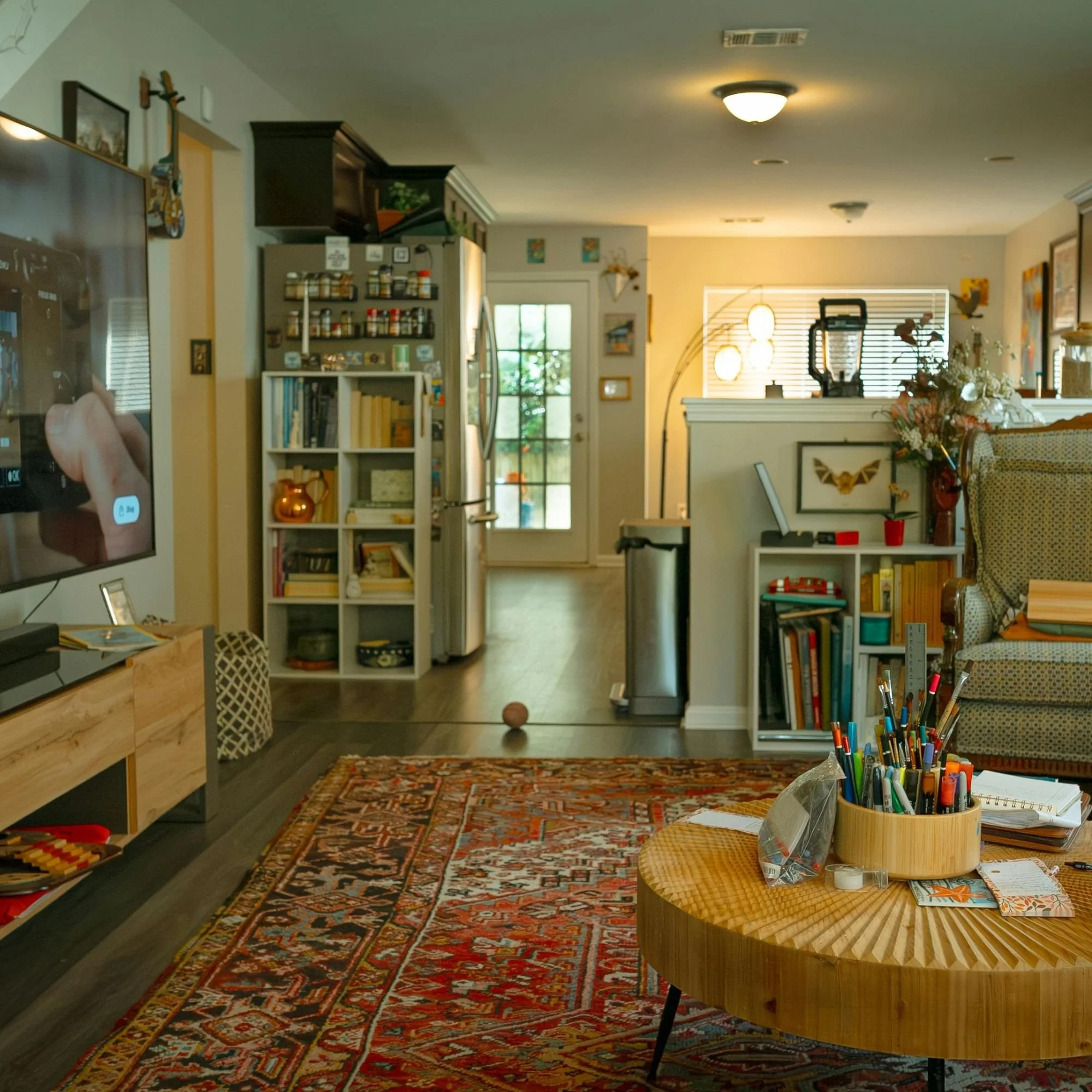 A cozy living room with a patterned area rug, surrounded by various furniture including a wooden table filled with pens and stationery, bookshelves, floral decorations, and wall art.