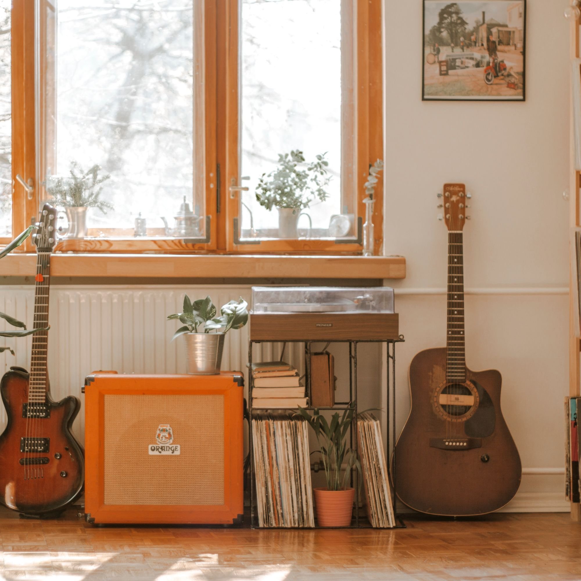 A view of a room with a large window showing snowy trees outside. Inside, there are two guitars, an orange guitar amp labeled "Orange," a table with a potted plant, a bookshelf filled with books and records, a framed painting on the wall, a guitar, an orange amplifier, a speaker, and a small wooden jewelry box.