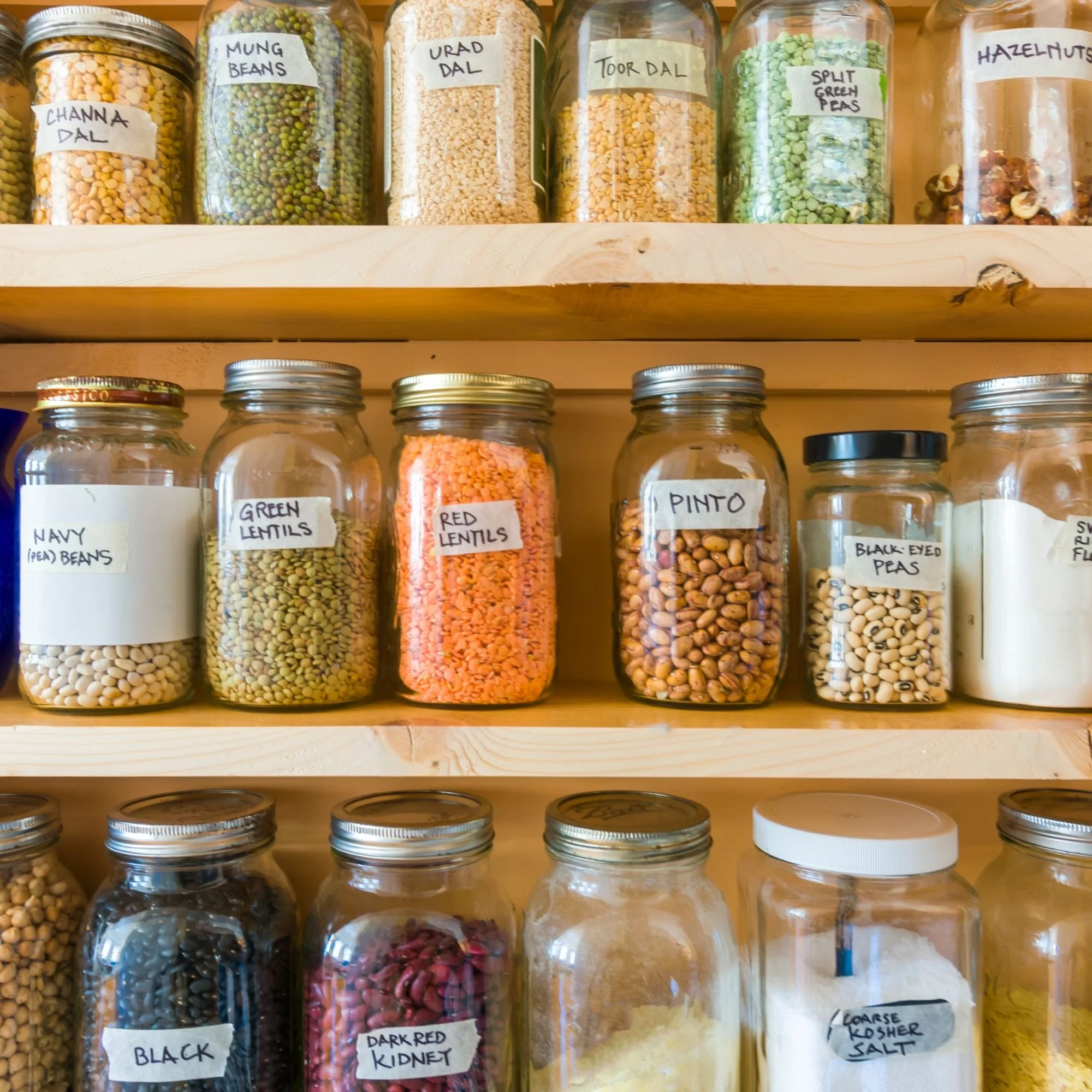 Shelf of glass jars filled with various dried legumes and grains, labeled with names such as mung beans, urad dal, toor dal, split green peas, hazelnuts, and other types of lentils and beans.