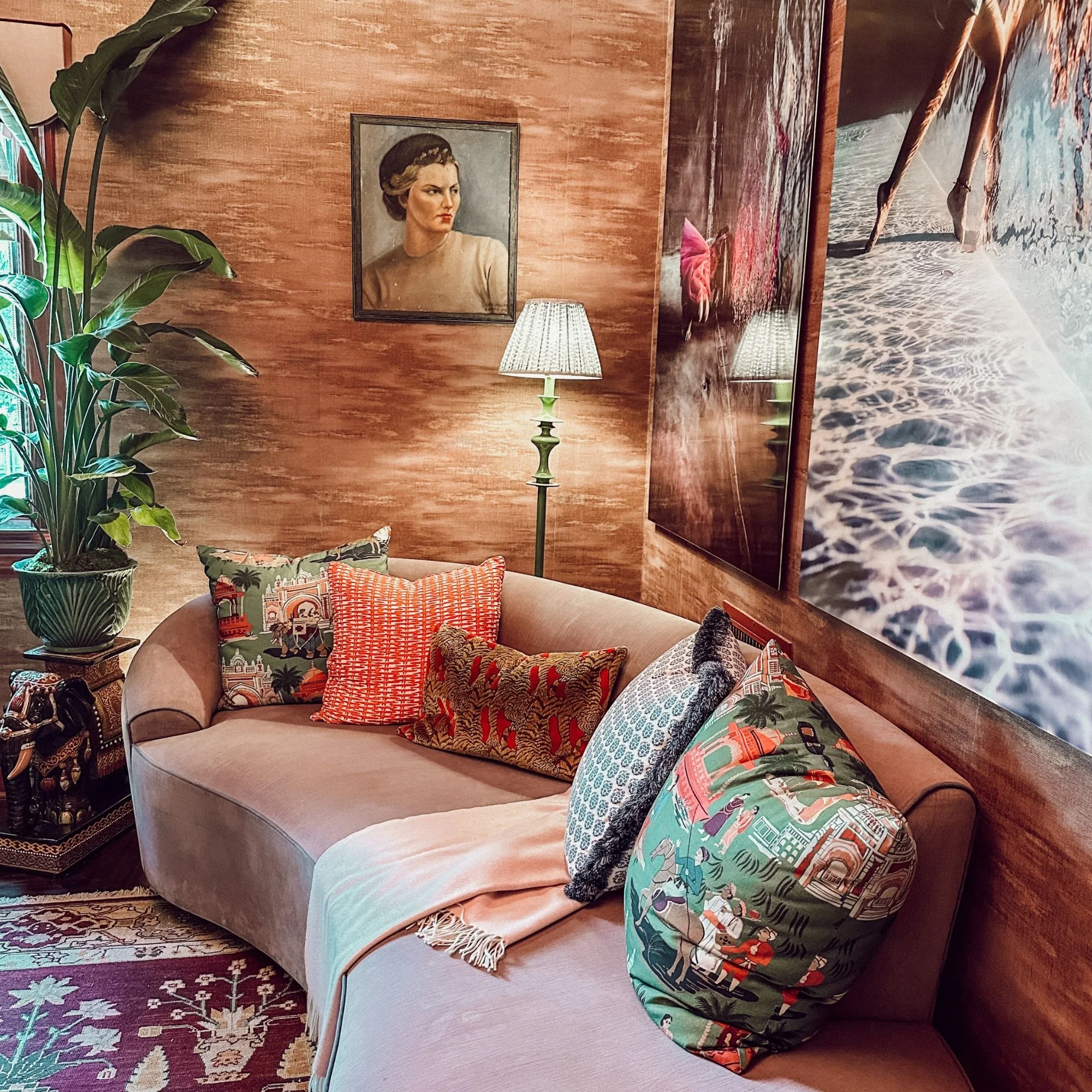 Living room with a beige sofa, decorative pillows, a large green plant, a floor lamp, and wall art including a portrait of a woman and a beach scene.