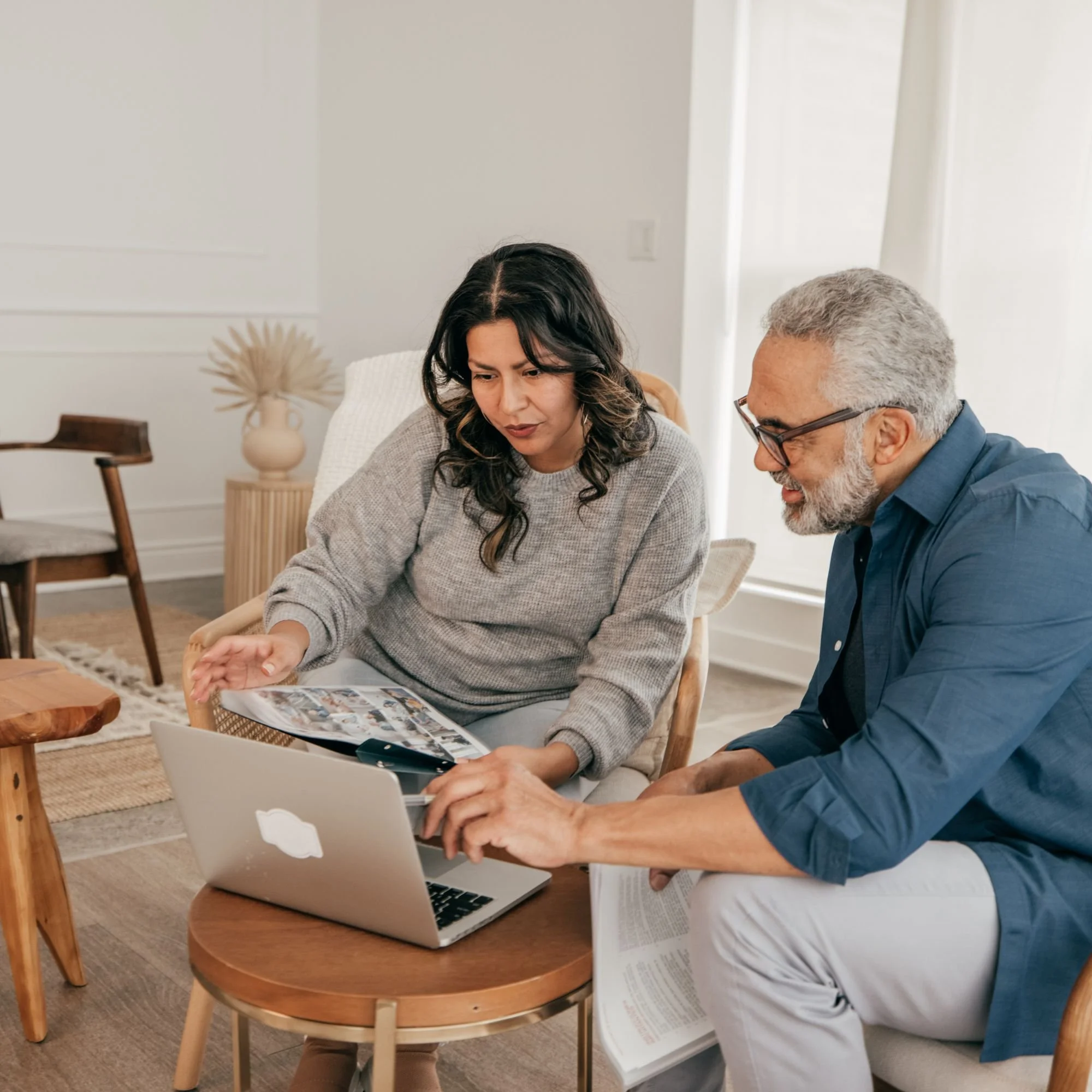 Two people, a woman with dark, wavy hair and a man with gray hair and beard, are sitting at a wooden table reviewing documents and using a laptop, in a bright, modern room.
