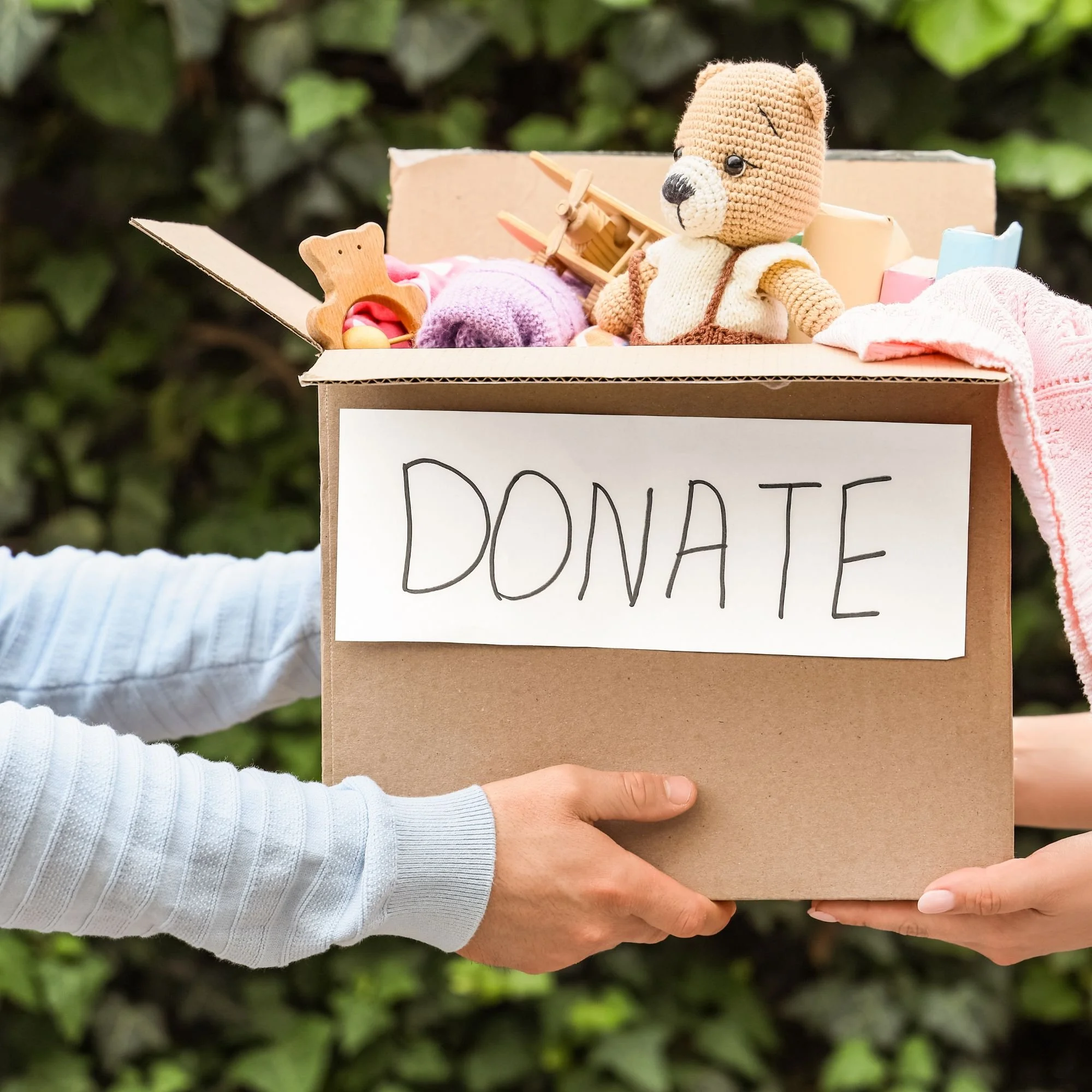 A person in a white long sleeve shirt holding a cardboard donation box filled with toys, including a knitted teddy bear, wooden toy airplane, and other soft toys, outdoors with green foliage in the background.