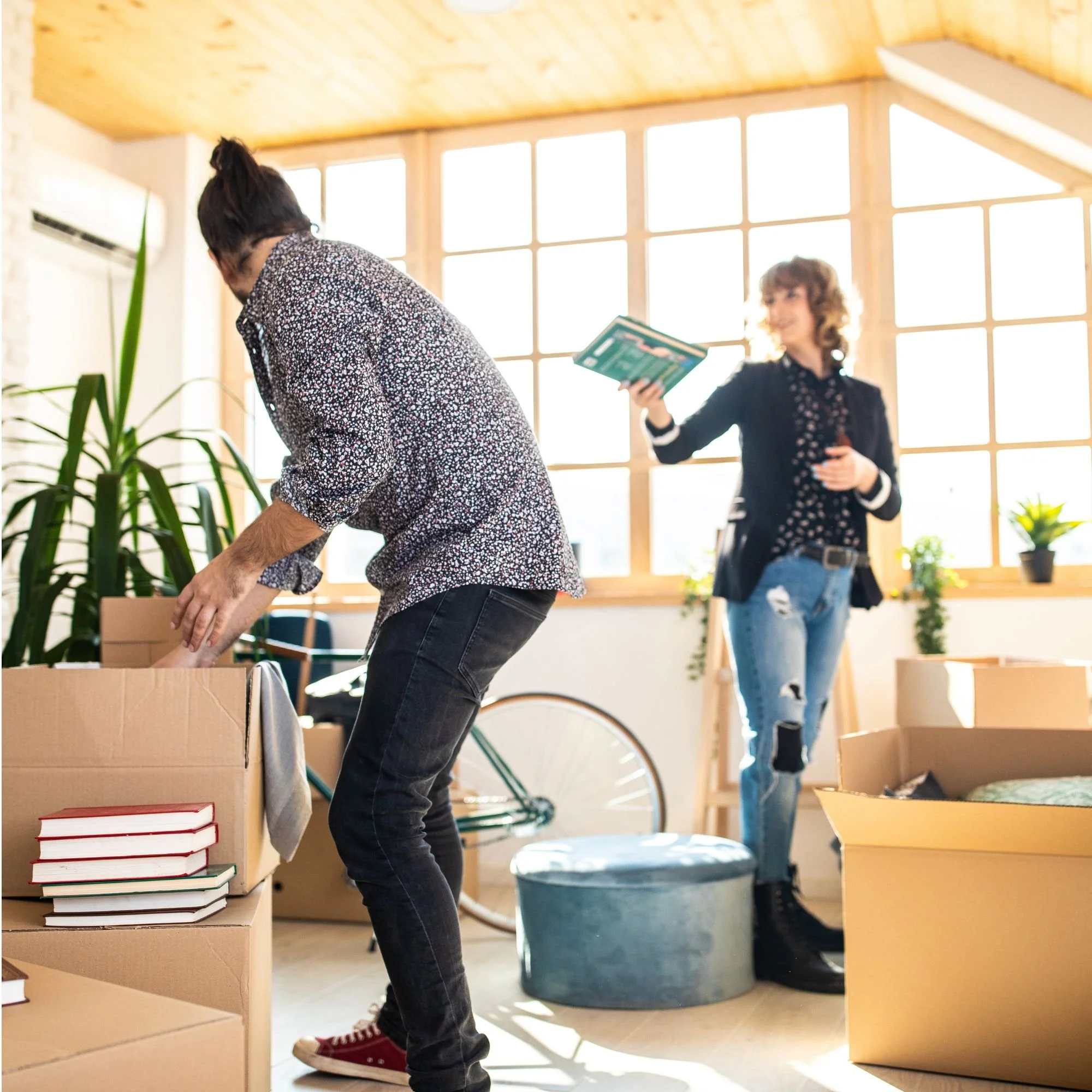 Two women packing books into cardboard boxes in a brightly lit room with large windows. One woman is wearing a patterned shirt and jeans, while the other is holding a book and wearing a dark jacket and ripped jeans.