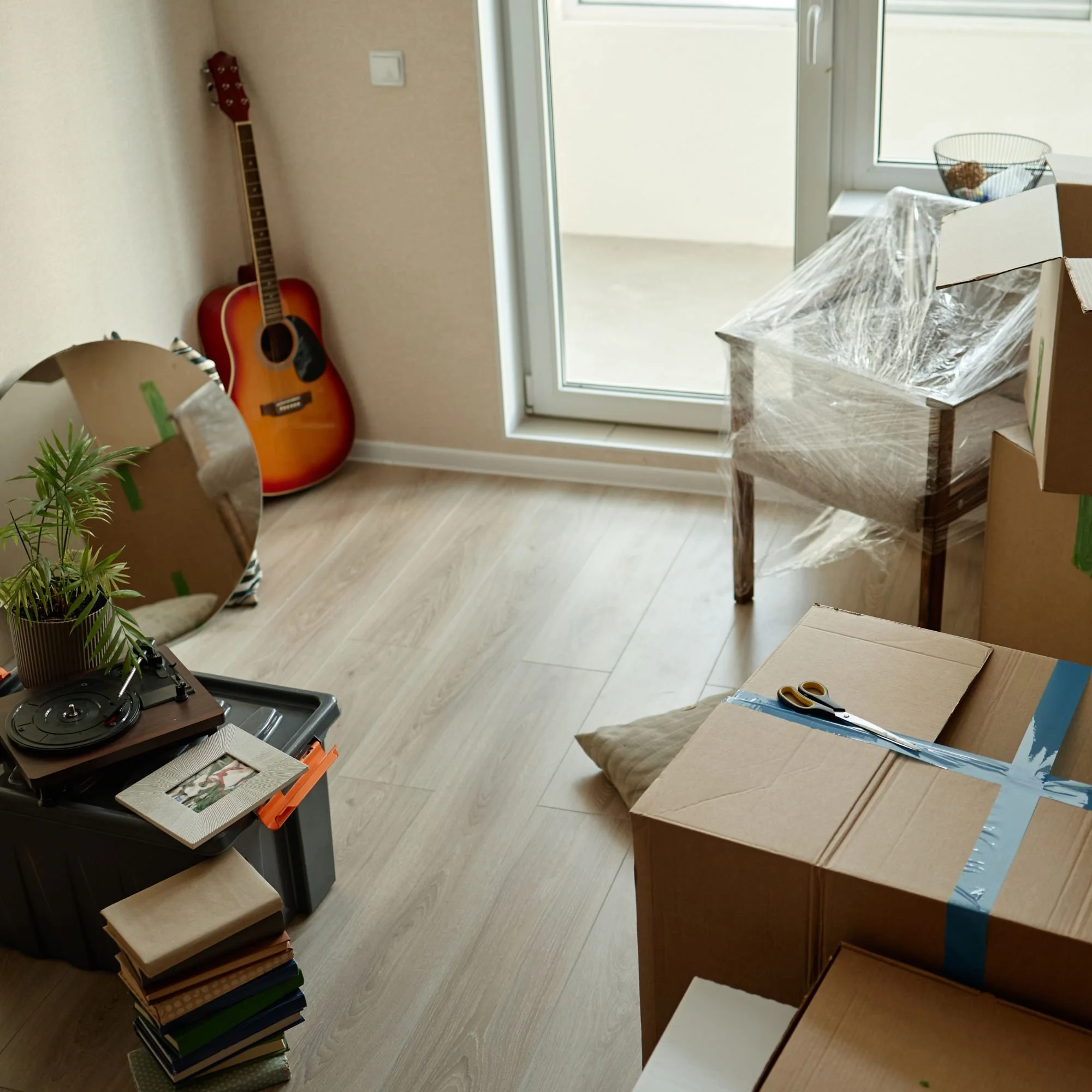 Room with a guitar leaning against the wall, a stack of books, a table with a cardboard box, a chair wrapped in plastic, and a sliding glass door leading to a balcony or patio.