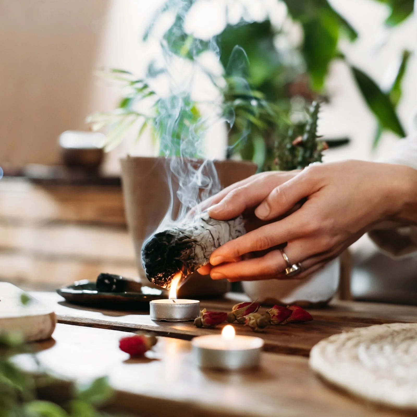 Person holding burning incense with a wooden table, candles, and potted plants in the background.