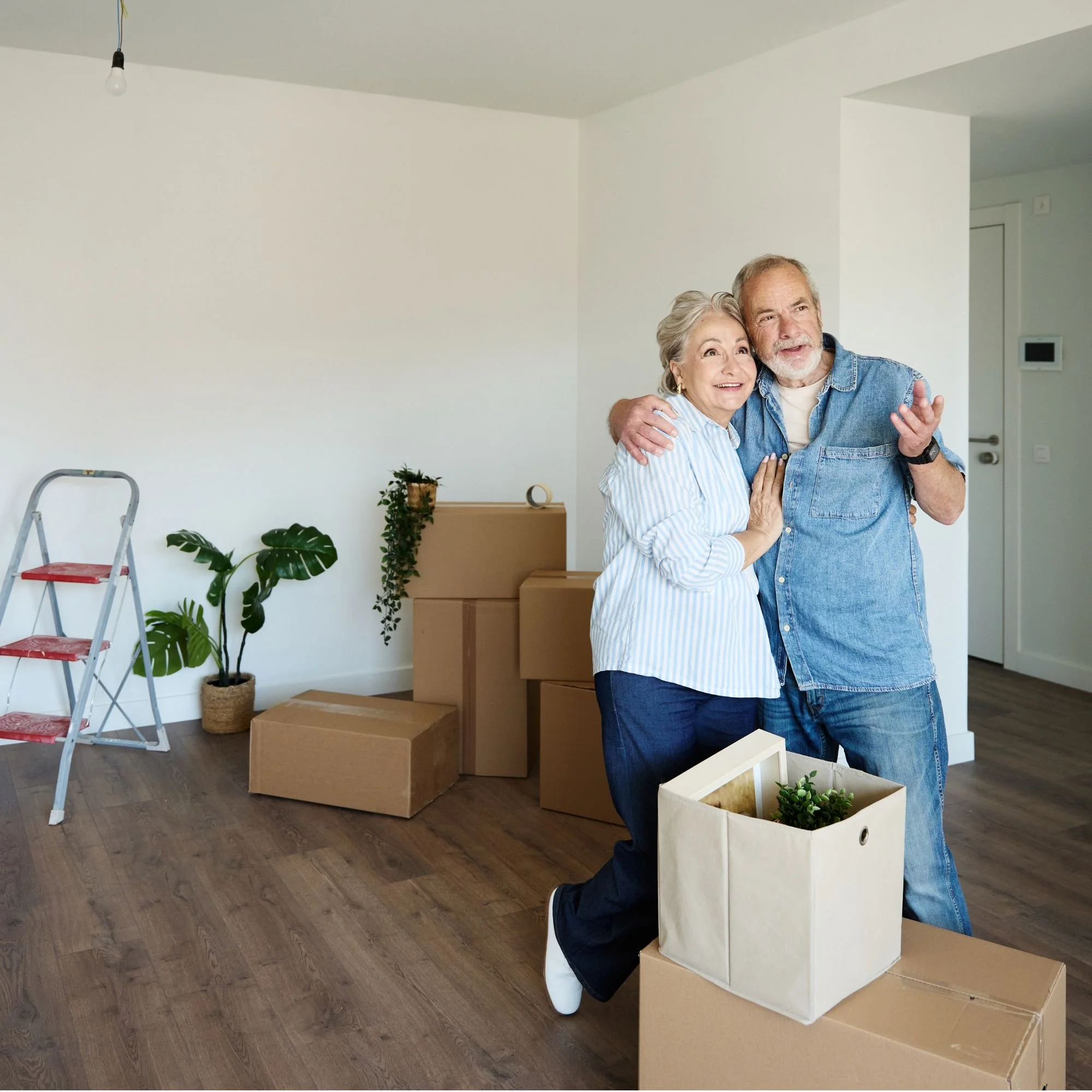 An elderly couple with gray hair, smiling and hugging while shopping for plants and home decor in a store. The man is wearing a denim shirt and jeans, the woman a light blue striped shirt. The store has wooden flooring, cardboard boxes, and plants, with an empty wall and some boxes in the background.