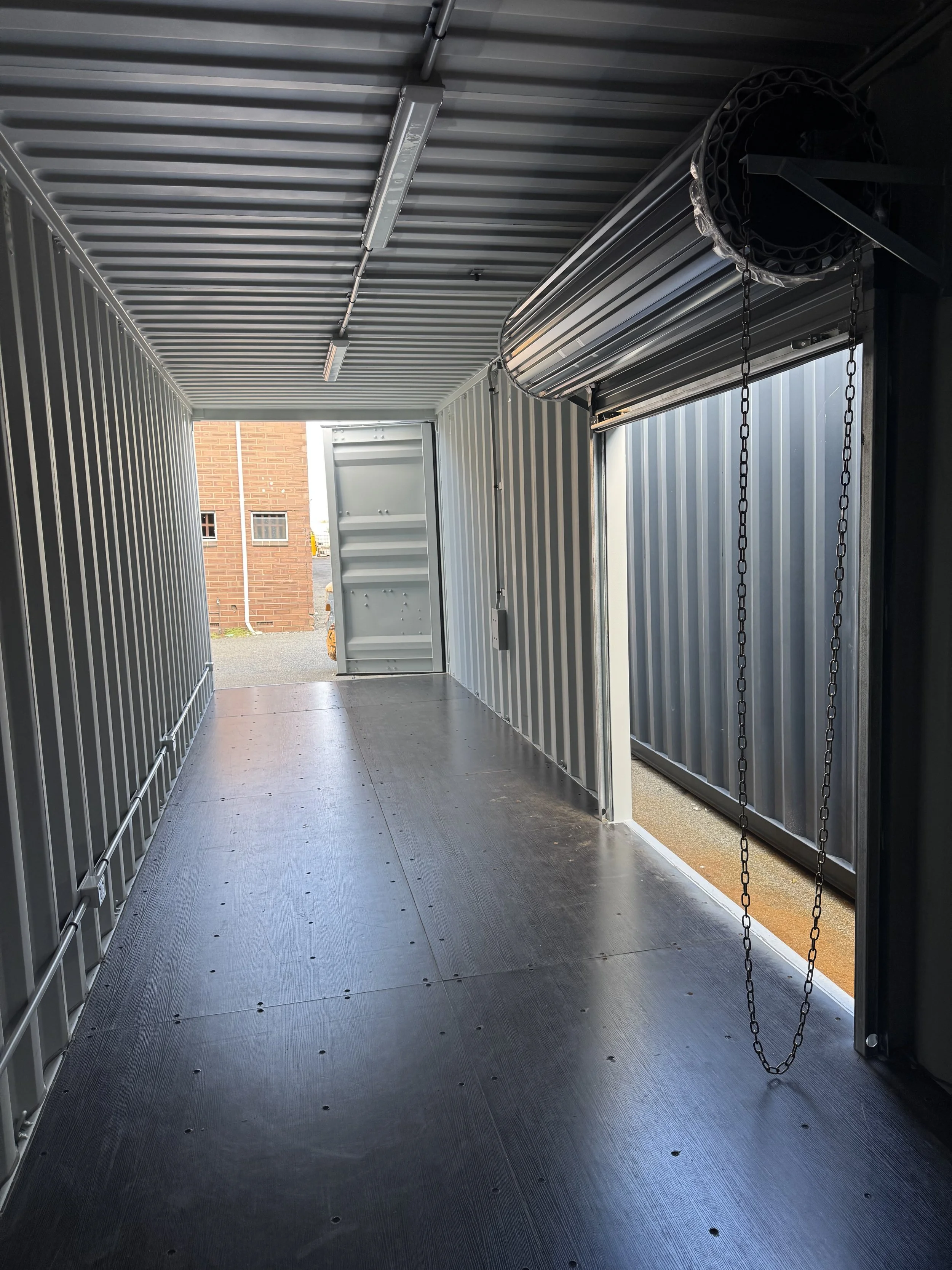 Interior of a cargo trailer with metal walls, a ribbed ceiling, and a rubber floor, showing an open door revealing a brick building outside.