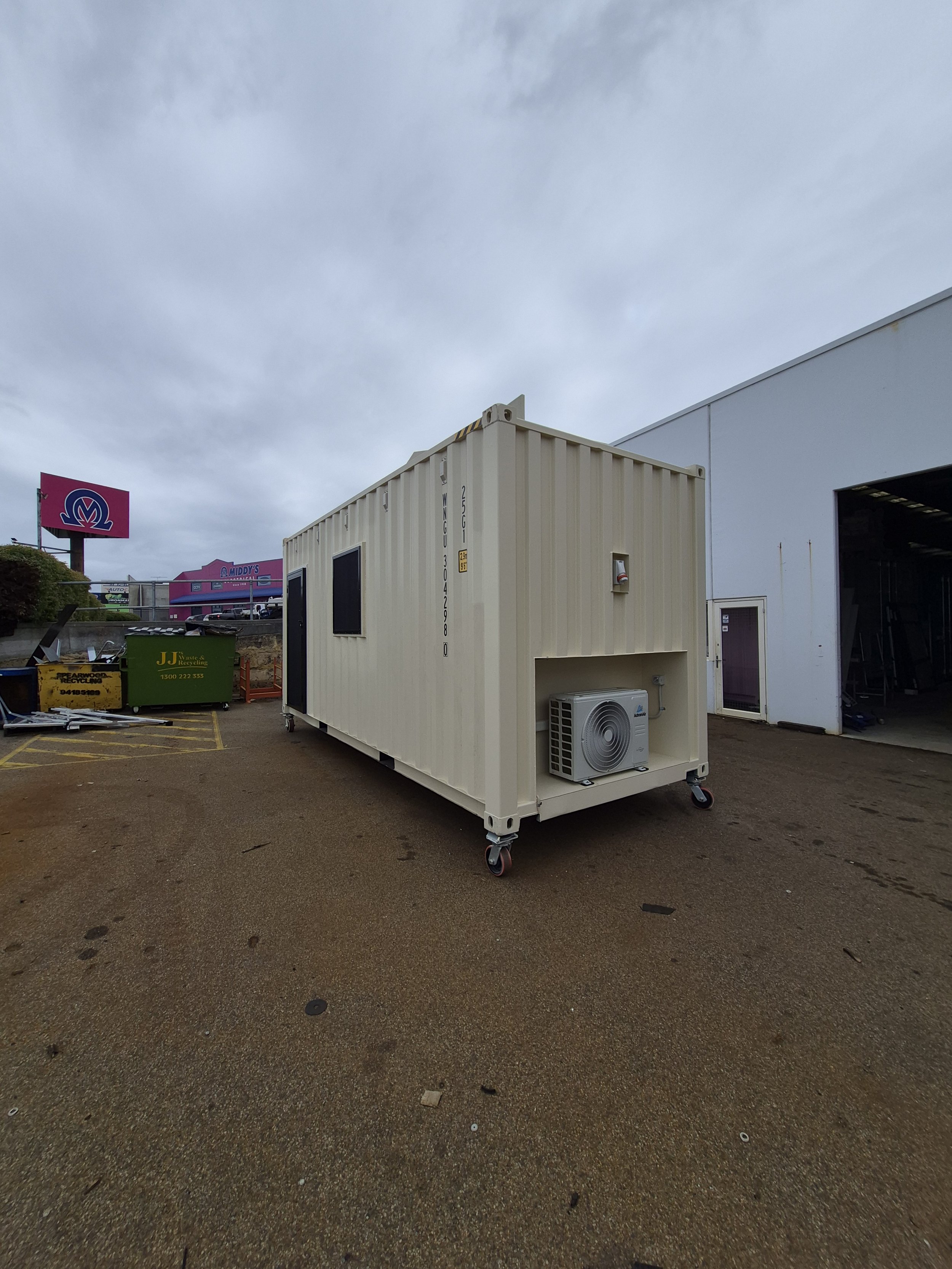 A beige shipping container on wheels with an air conditioning unit outside, located in a parking lot with trash bins and a partly open warehouse. Overcast sky overhead.