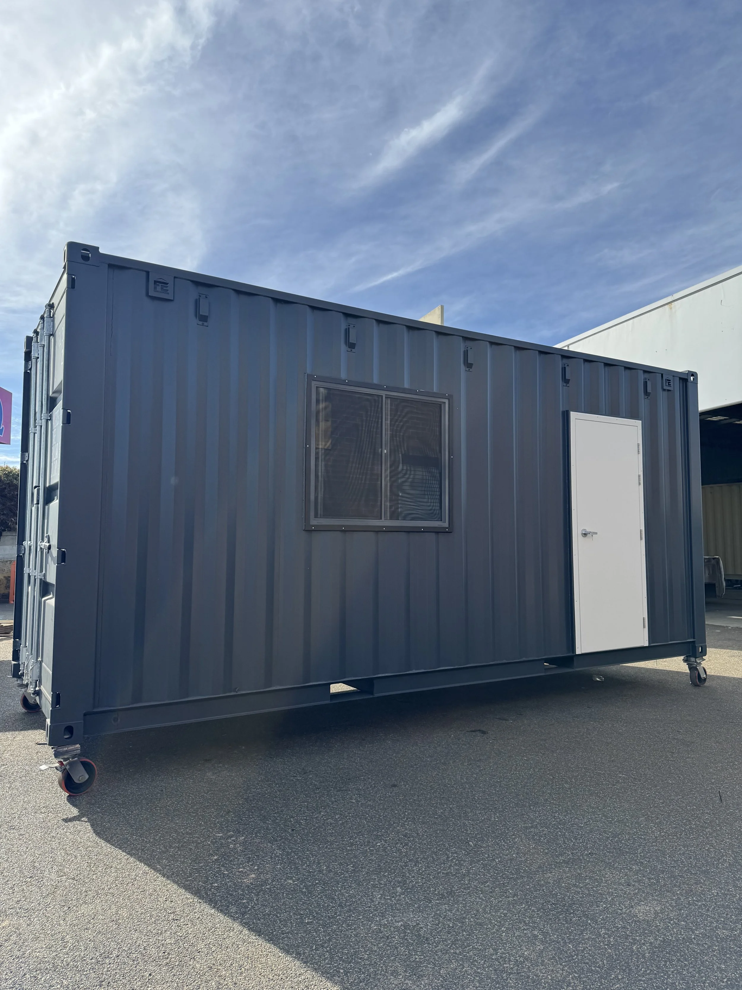 A gray portable container on wheels with a window and a door, located outdoors against a blue sky with some clouds.