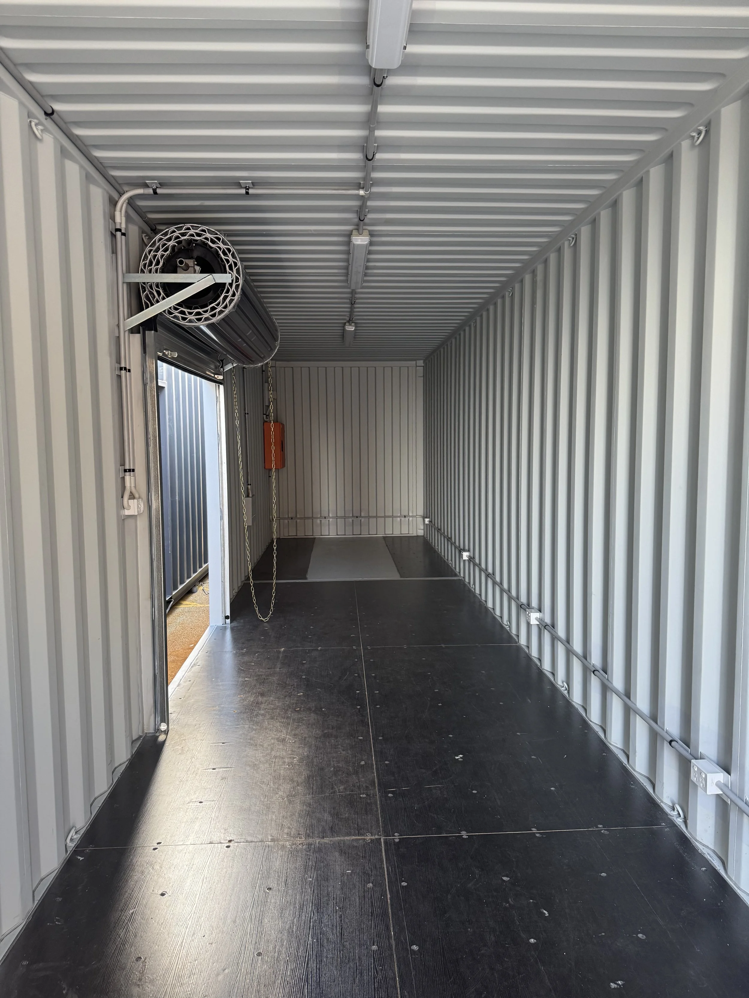 Empty cargo container interior with metal walls, ceiling, and floor, with electrical fixtures along the top and wall, and chains hanging from the ceiling.
