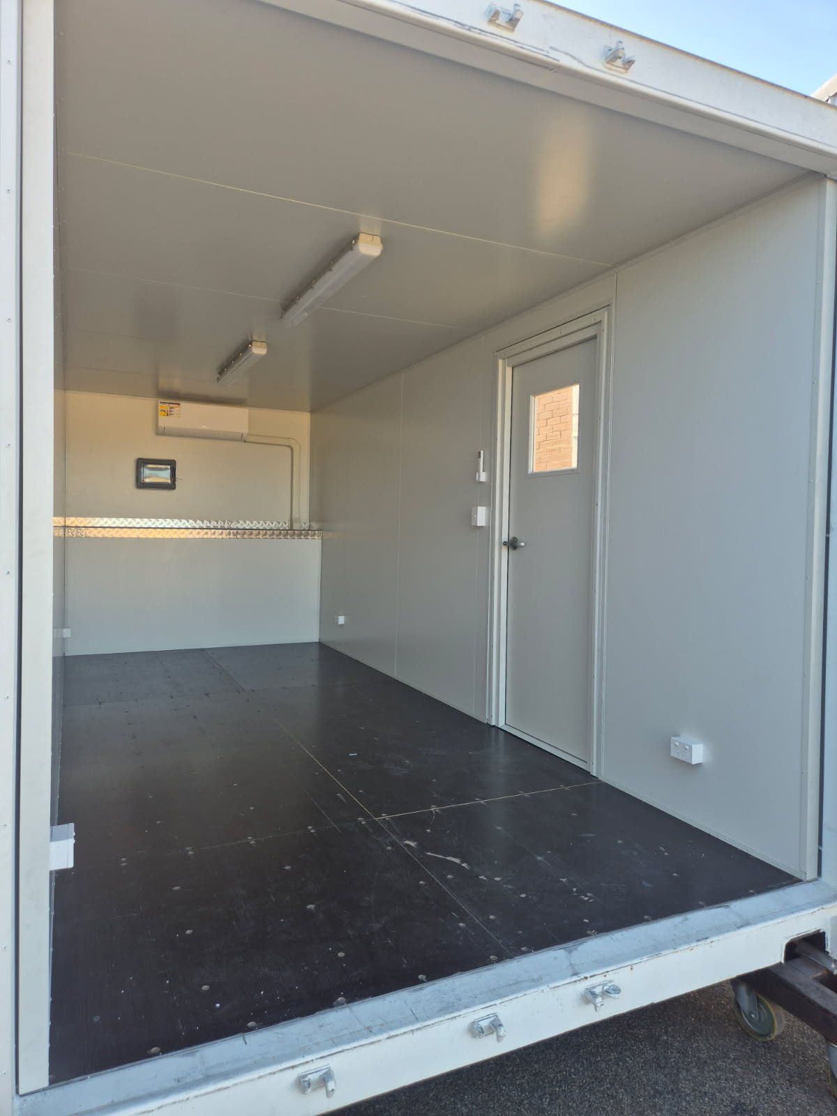 Empty mobile workspace with gray interior walls, black floor, and a small windowed door, ceiling-mounted fluorescent lights, and a wall-mounted climate control unit.
