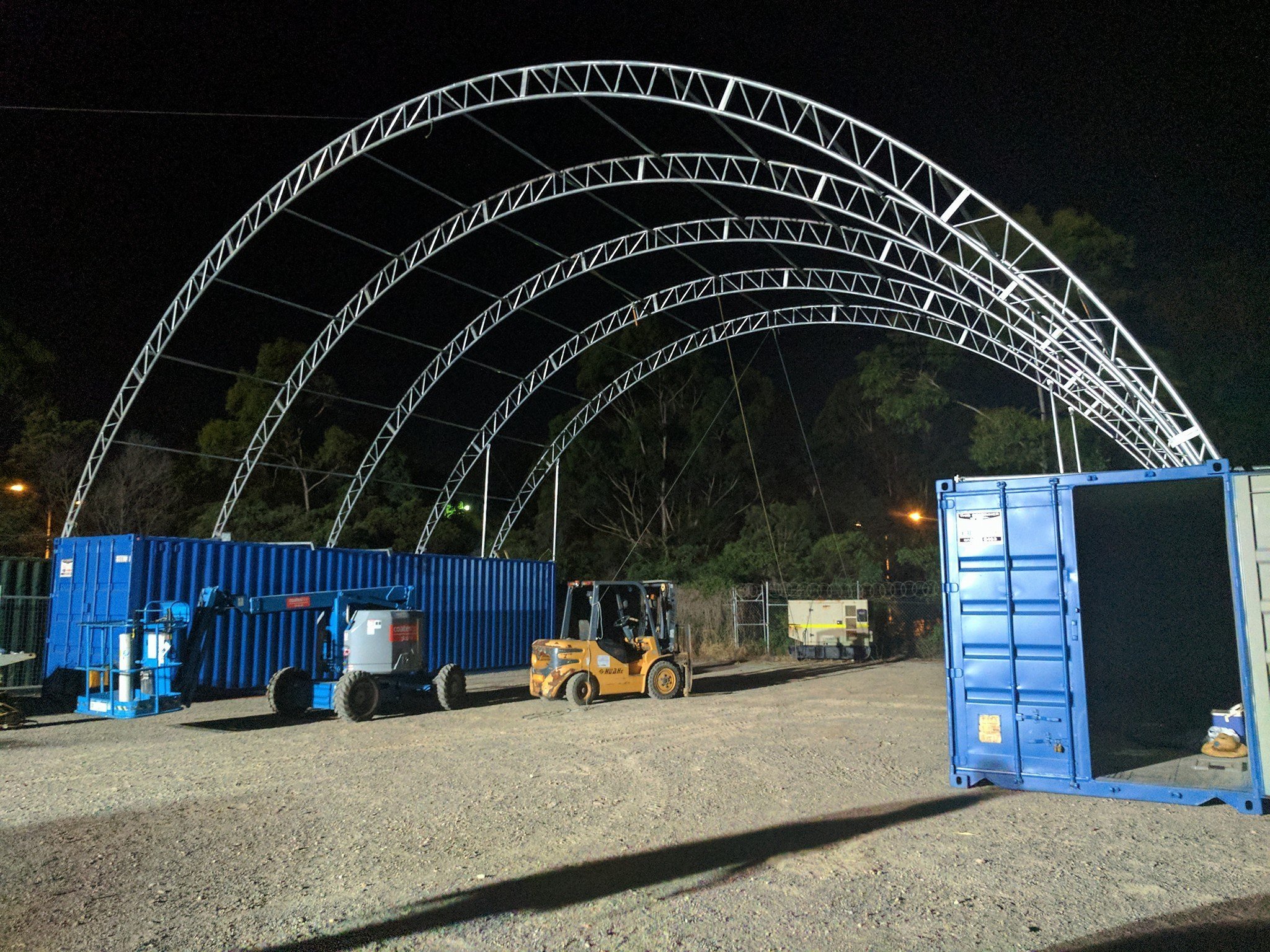 Nighttime construction site with metal arches being assembled, blue containers, a forklift, and a gravel ground.