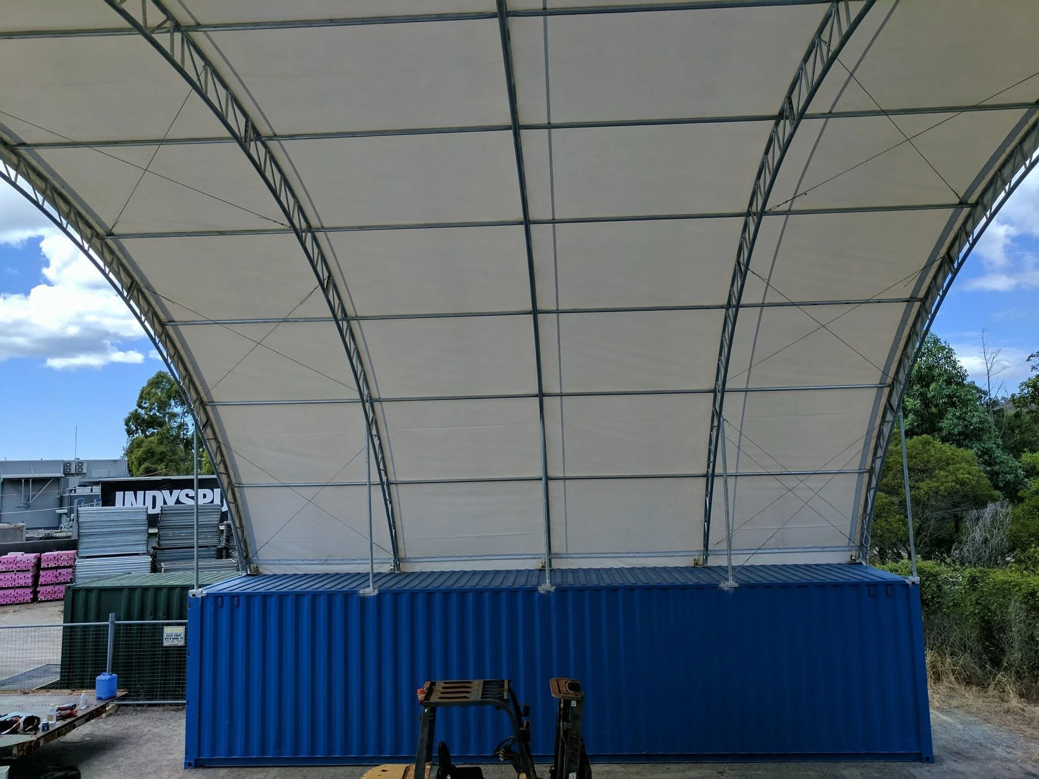 Large white outdoor stage tent with metal framework, blue shipping container structure underneath, and trees and sky in the background.