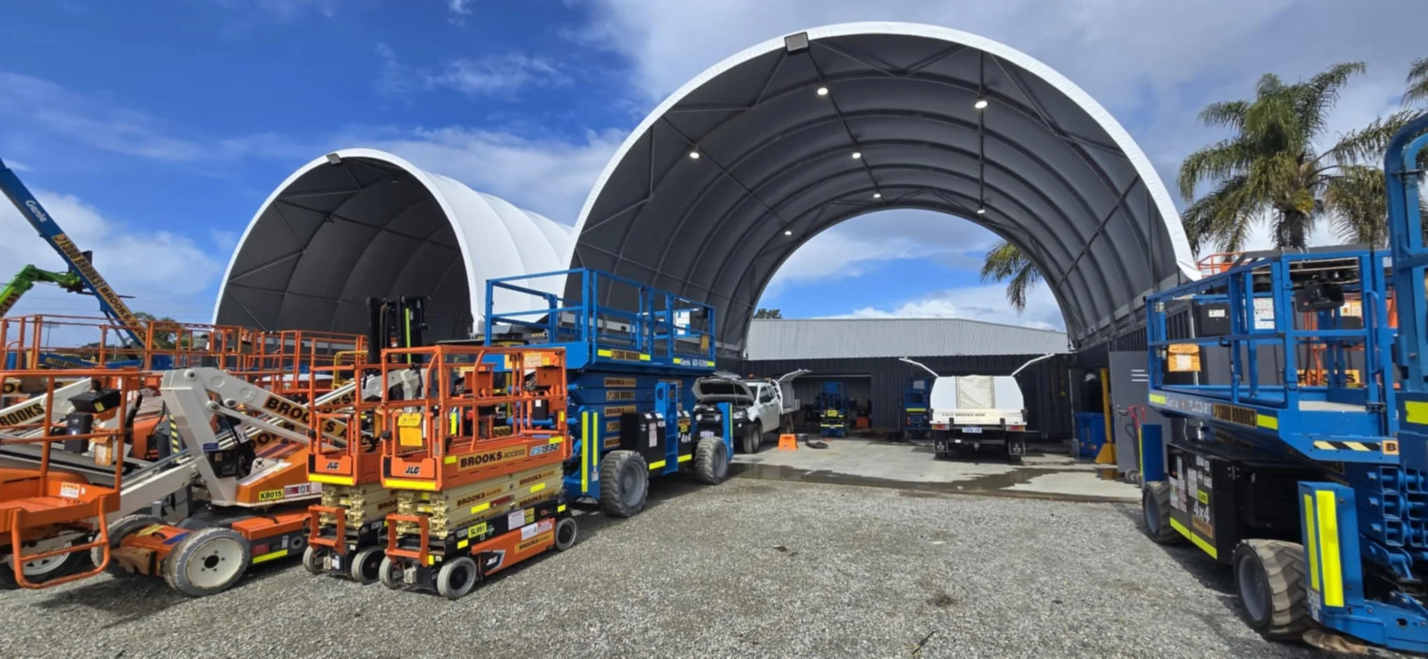 Construction site with large curved metal structures, lift equipment, and trucks under a blue sky with clouds and palm trees.