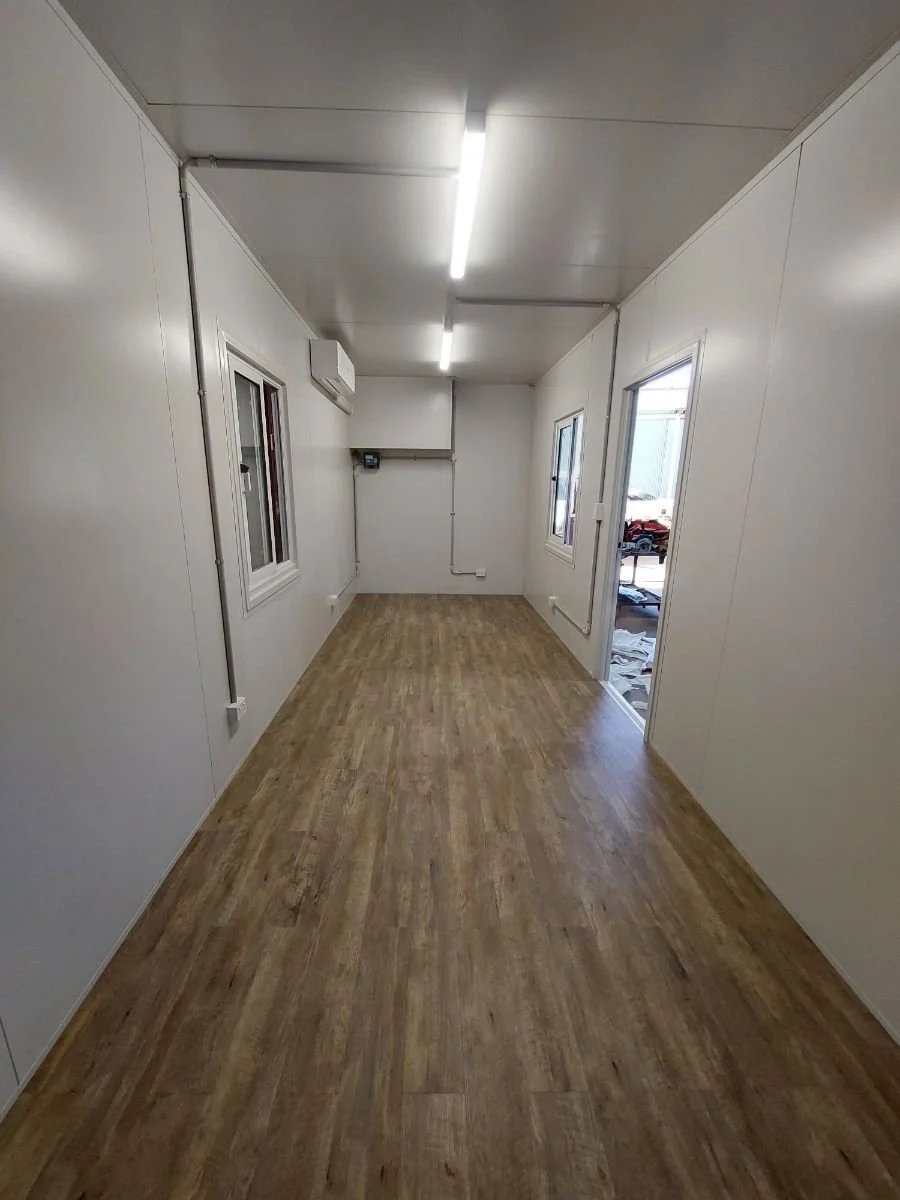 Empty rectangular room with wood flooring, white walls, and ceiling with fluorescent lights, contains windows and a door, some construction materials outside.