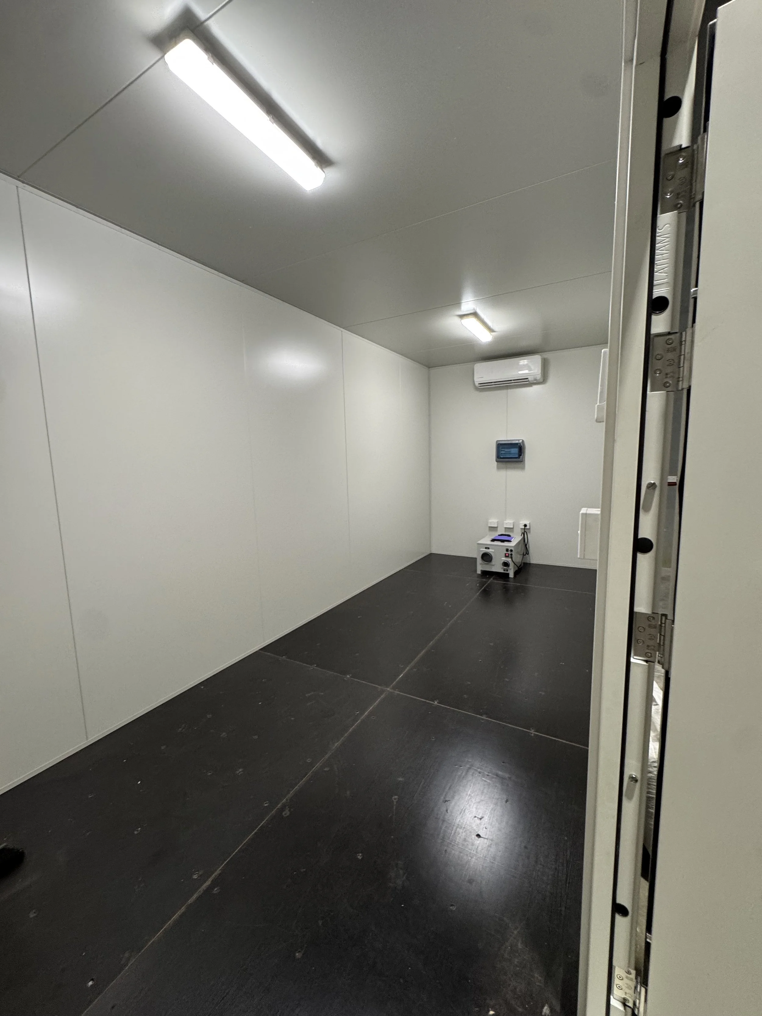 The image shows a small, empty, windowless room with white walls, black flooring, and fluorescent ceiling lights. There is a small heater or appliance on the floor and a control panel on the wall.