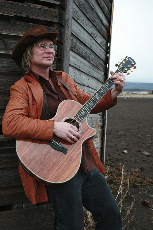 A man in country-style clothing, wearing a cowboy hat, glasses, and a leather jacket, standing outdoors near a wooden barn, playing an acoustic guitar.