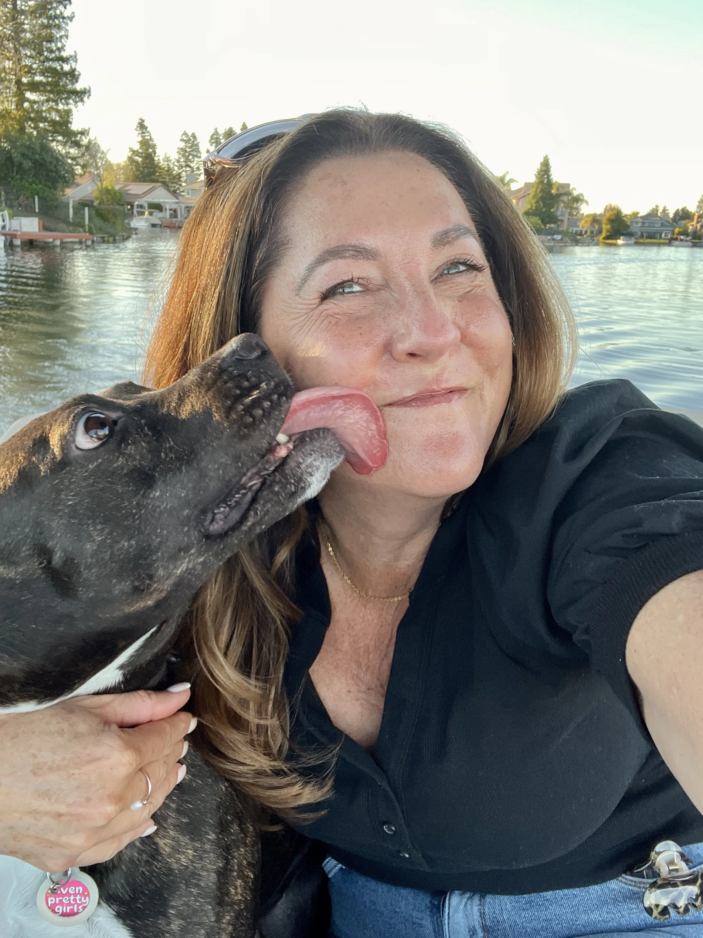 A woman with brown hair taking a selfie with a black dog on her lap, both near a body of water with houses and trees in the background. The woman is smiling and the dog looks up at her, licking her face.