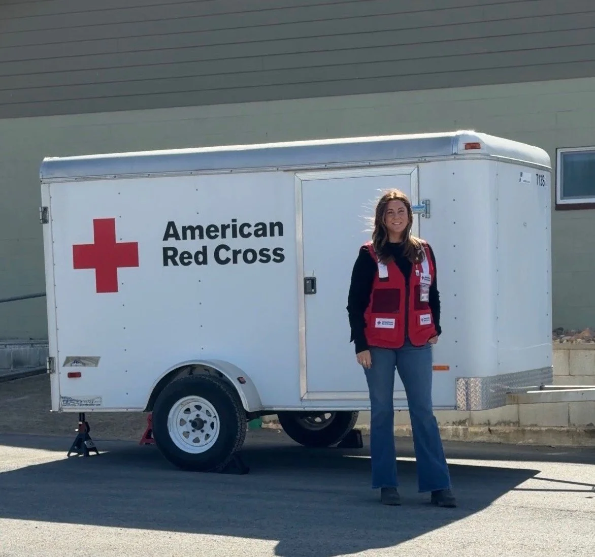 Woman smiling, standing outside next to a trailer with American Red Cross logo and text, wearing a red vest with white labels.