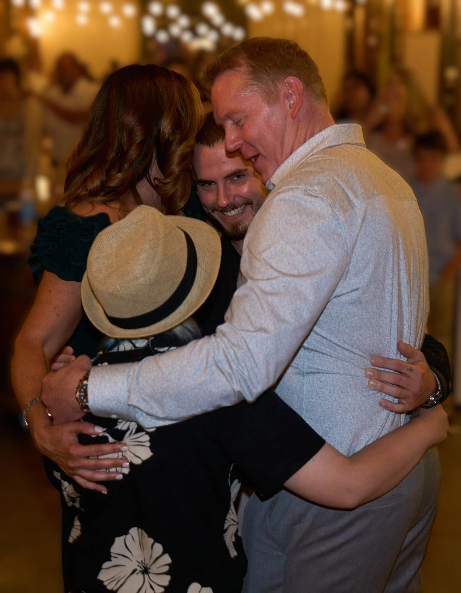 Three people hugging closely at a social gathering, with warm lighting in the background.