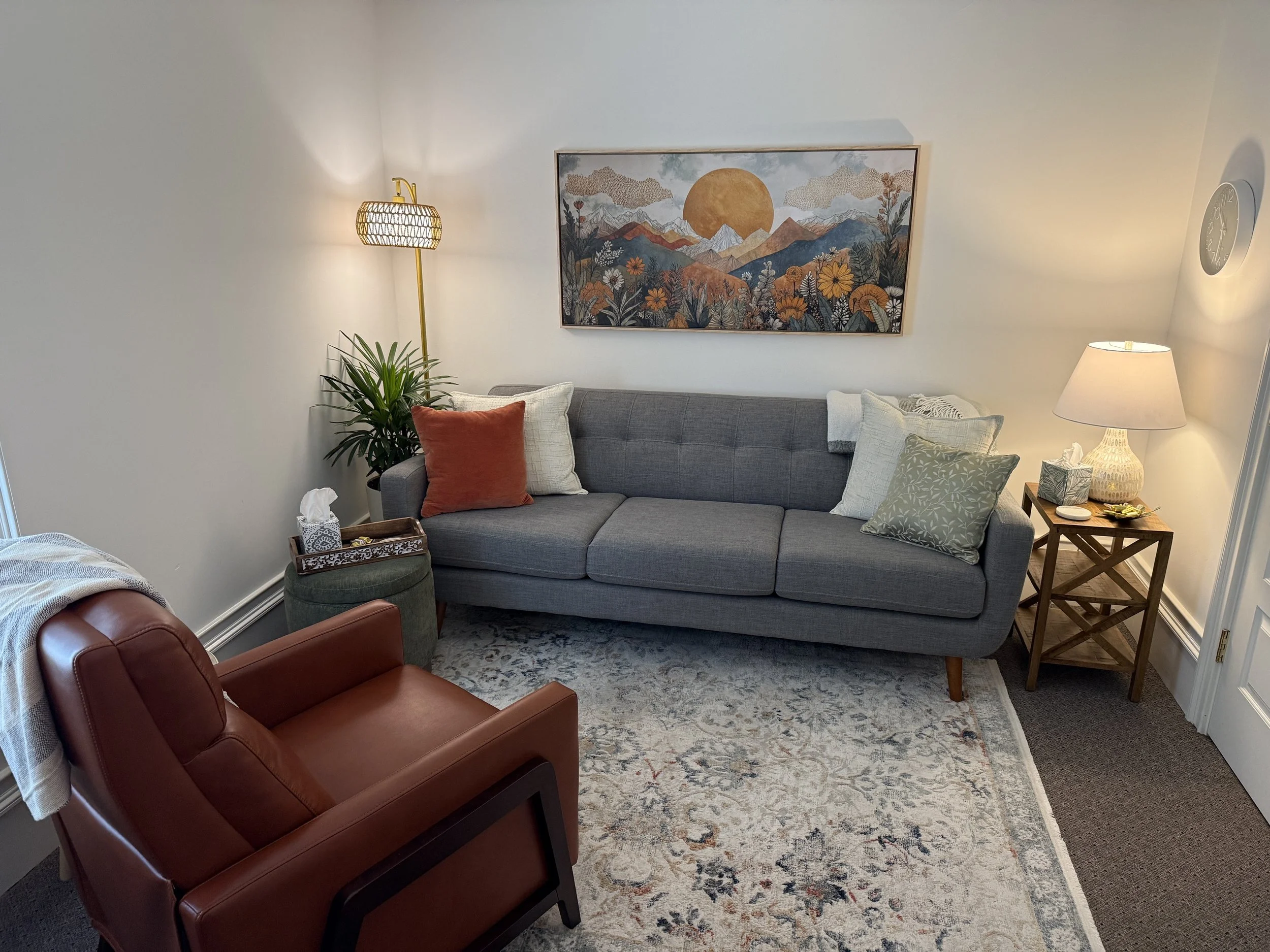 A cozy living room with a gray sofa adorned with various pillows, a wooden side table with a lamp, a tall plant, and framed scenic artwork on the wall. A leather armchair and a patterned rug are also visible.