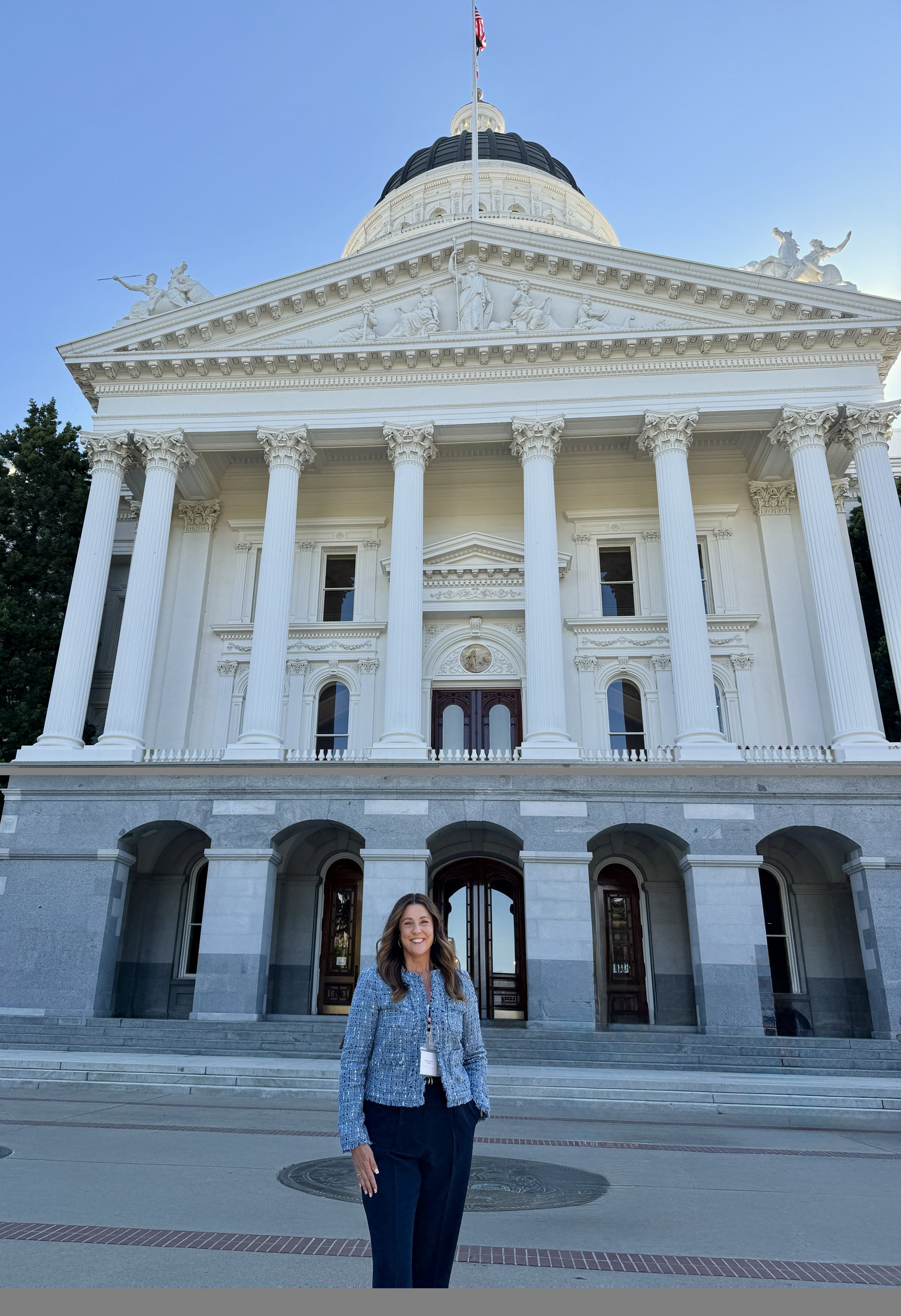 A woman standing in front of the Alabama State Capitol building, which features white columns, staircase, and detailed sculptures on the balustrade, with a dome and flag overhead, on a clear day.