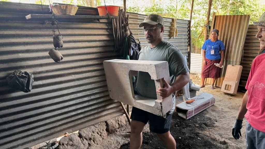 A man carrying a concrete microwave oven in a makeshift outdoor setting with corrugated metal walls, while two women observe. The area appears to be a workshop or construction site.
