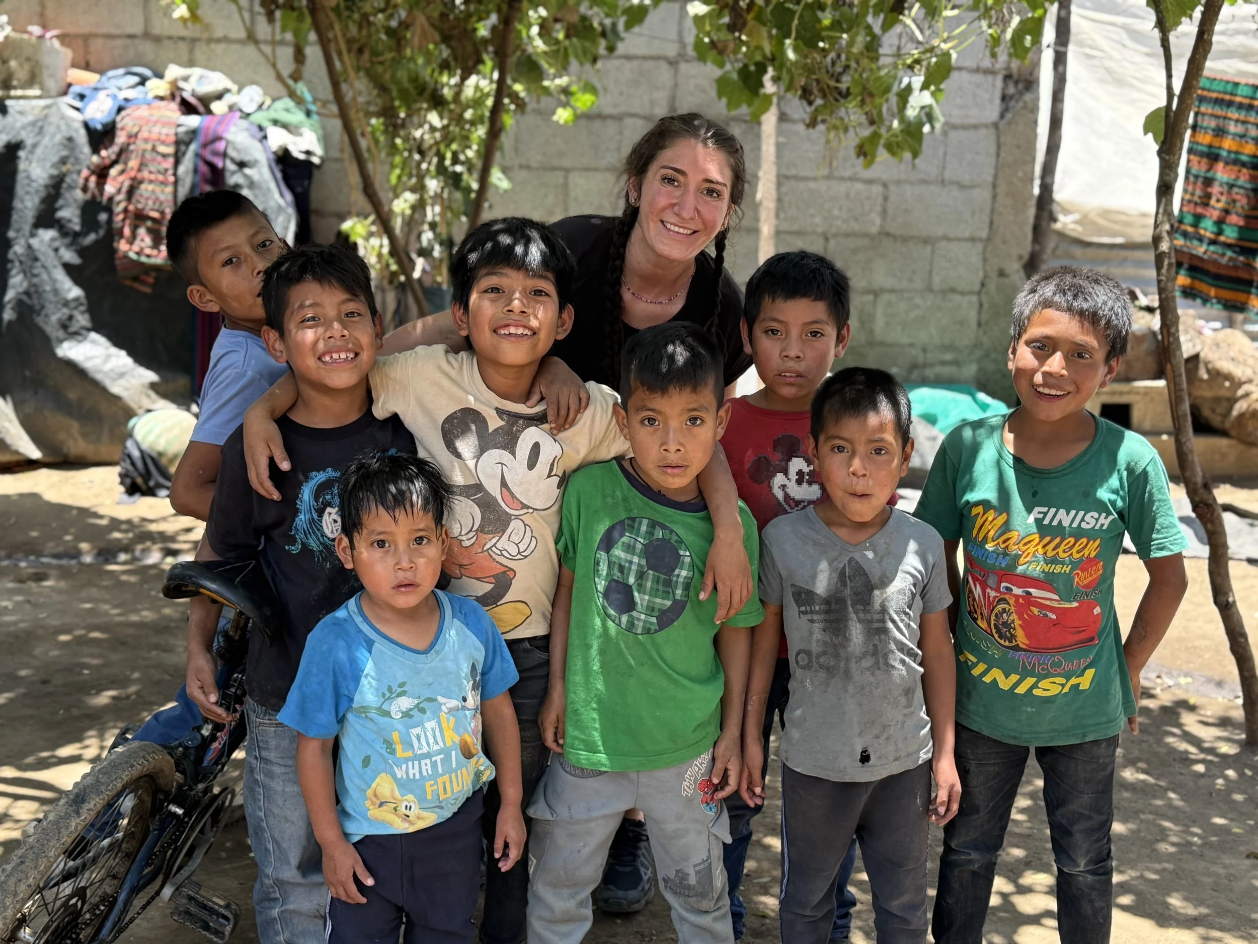 A smiling young woman with brown hair in braids standing among a group of diverse children outdoors, with a brick wall, trees, and laundry hanging in the background.