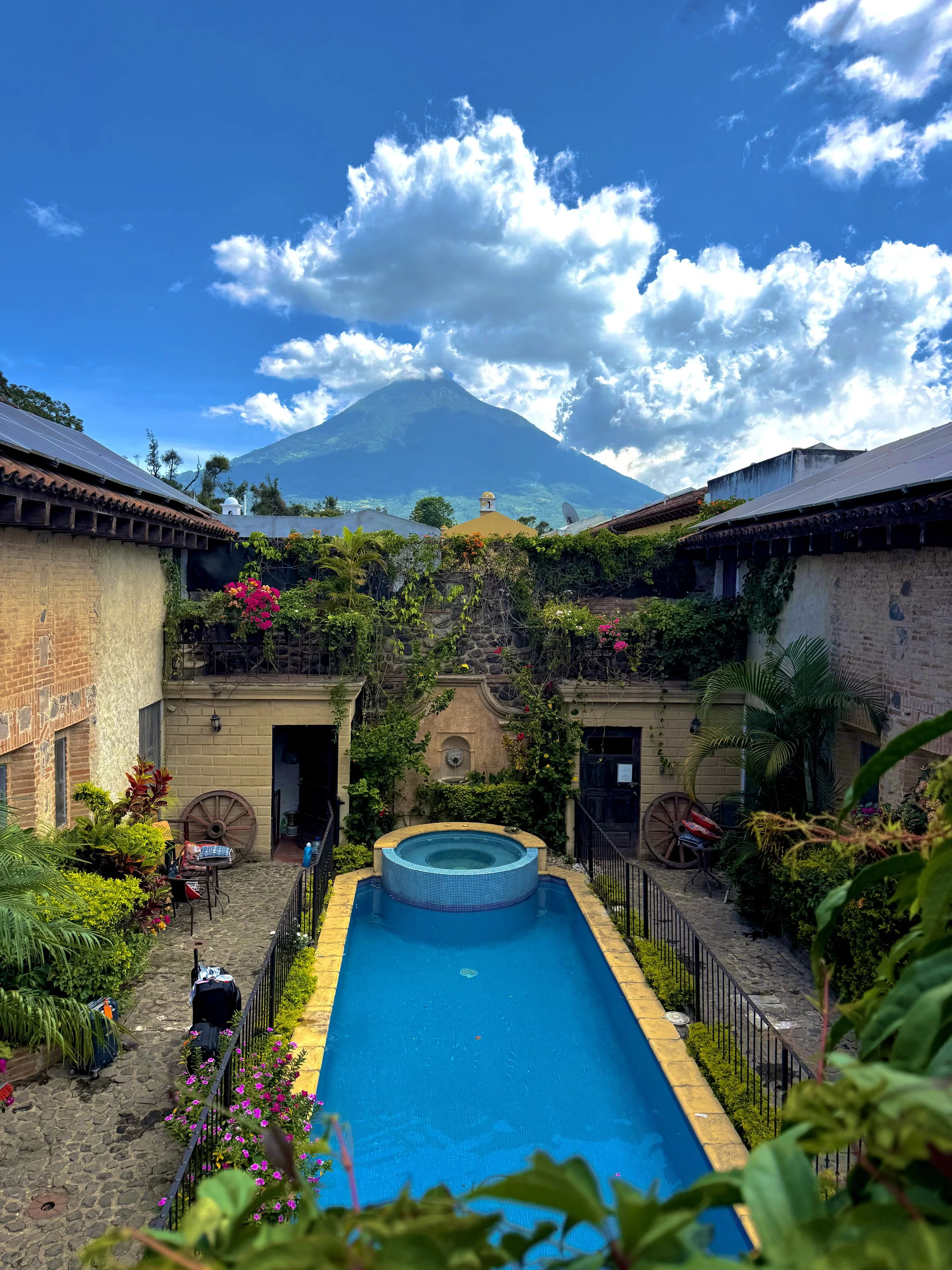 A backyard pool with clear blue water, surrounded by stone paving and lush green plants and flowers, with a mountain in the background and a partly cloudy sky.