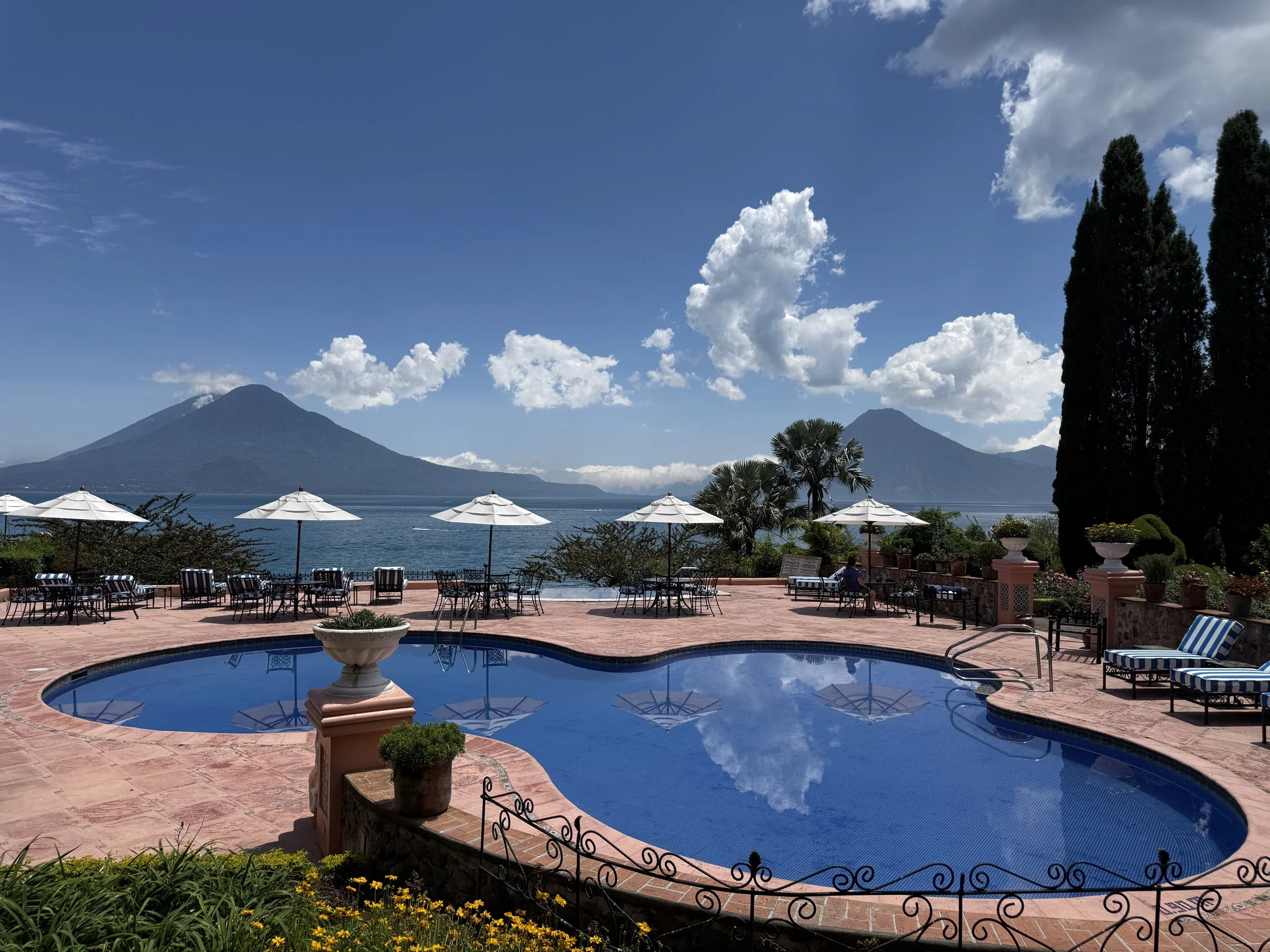 A scenic view of a swimming pool area with umbrellas, chairs, and potted plants, overlooking a large lake with mountains in the background and a partly cloudy sky.