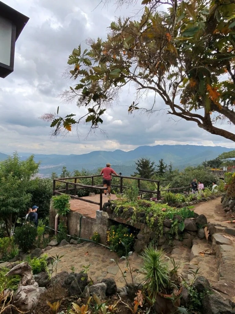 People enjoying a scenic view from a garden lookout on a mountain, surrounded by lush greenery and overcast skies.