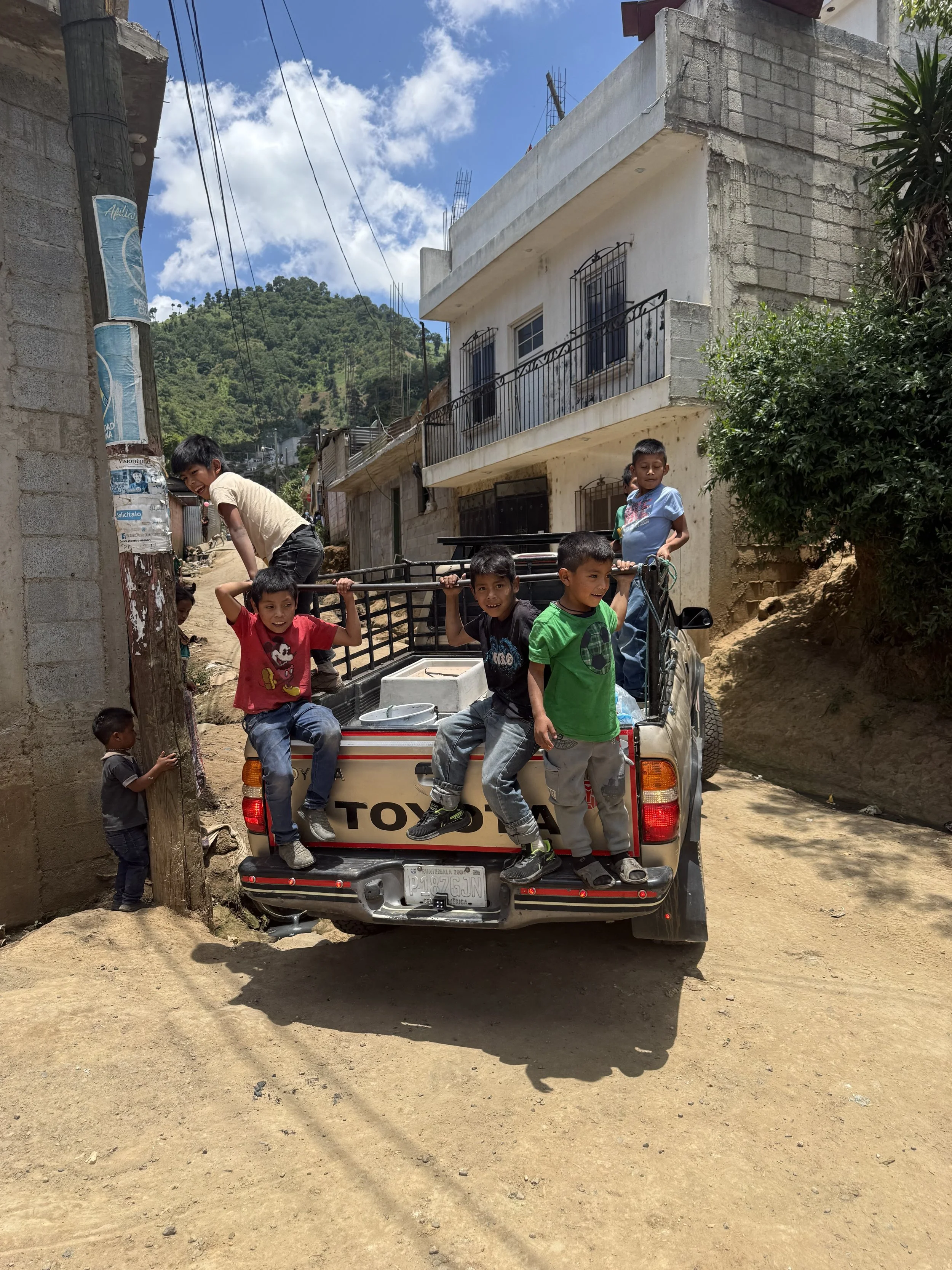 A group of children riding and playing on the back of a small pickup truck on a dirt road in a rural neighborhood. The neighborhood has some unfinished and finished houses, with mountains and a partly cloudy sky in the background.