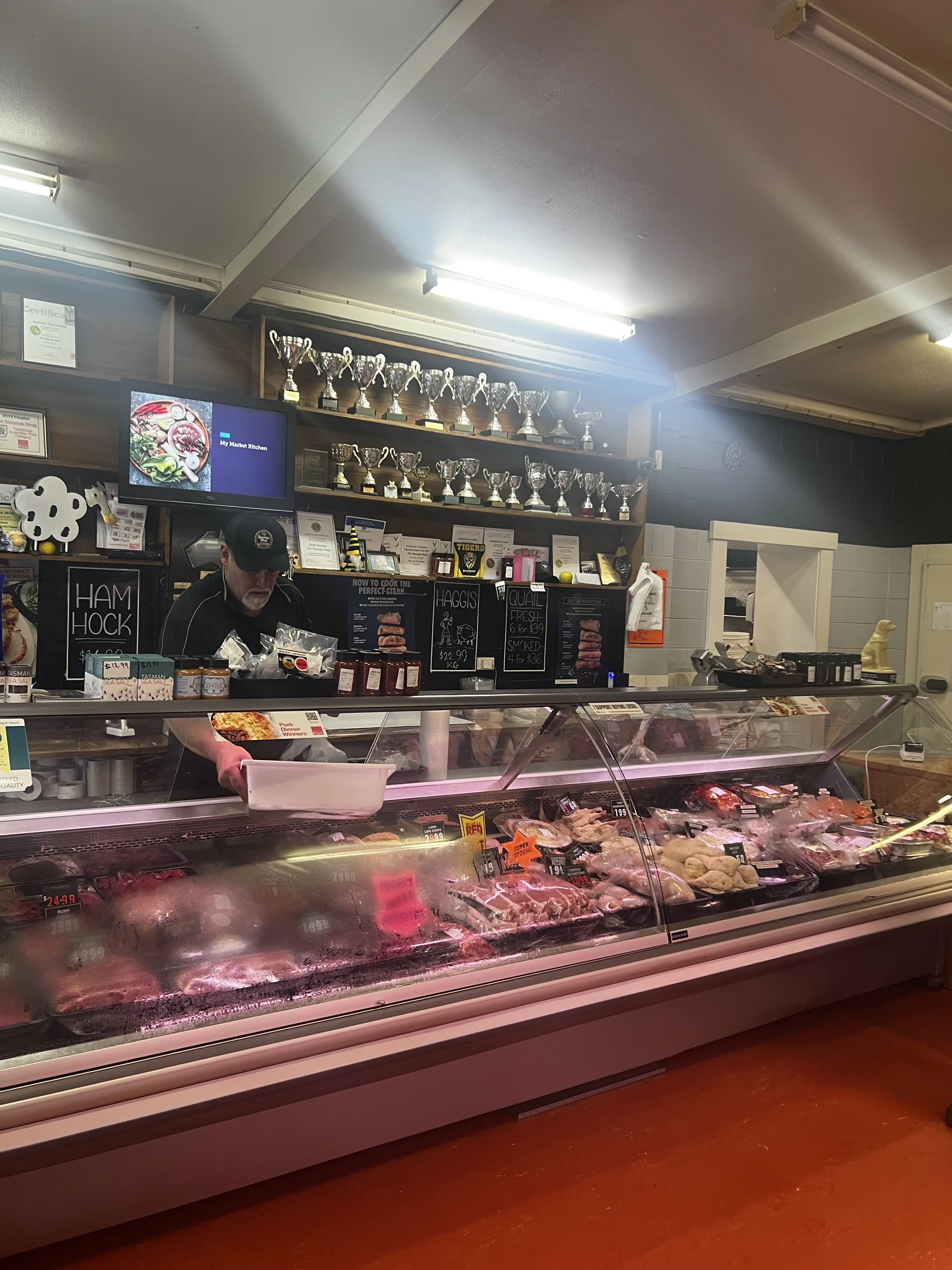 Display case with various cuts of meat and seafood in a grocery store, with a worker behind the counter. Trophies and certificates are visible on the shelves above.
