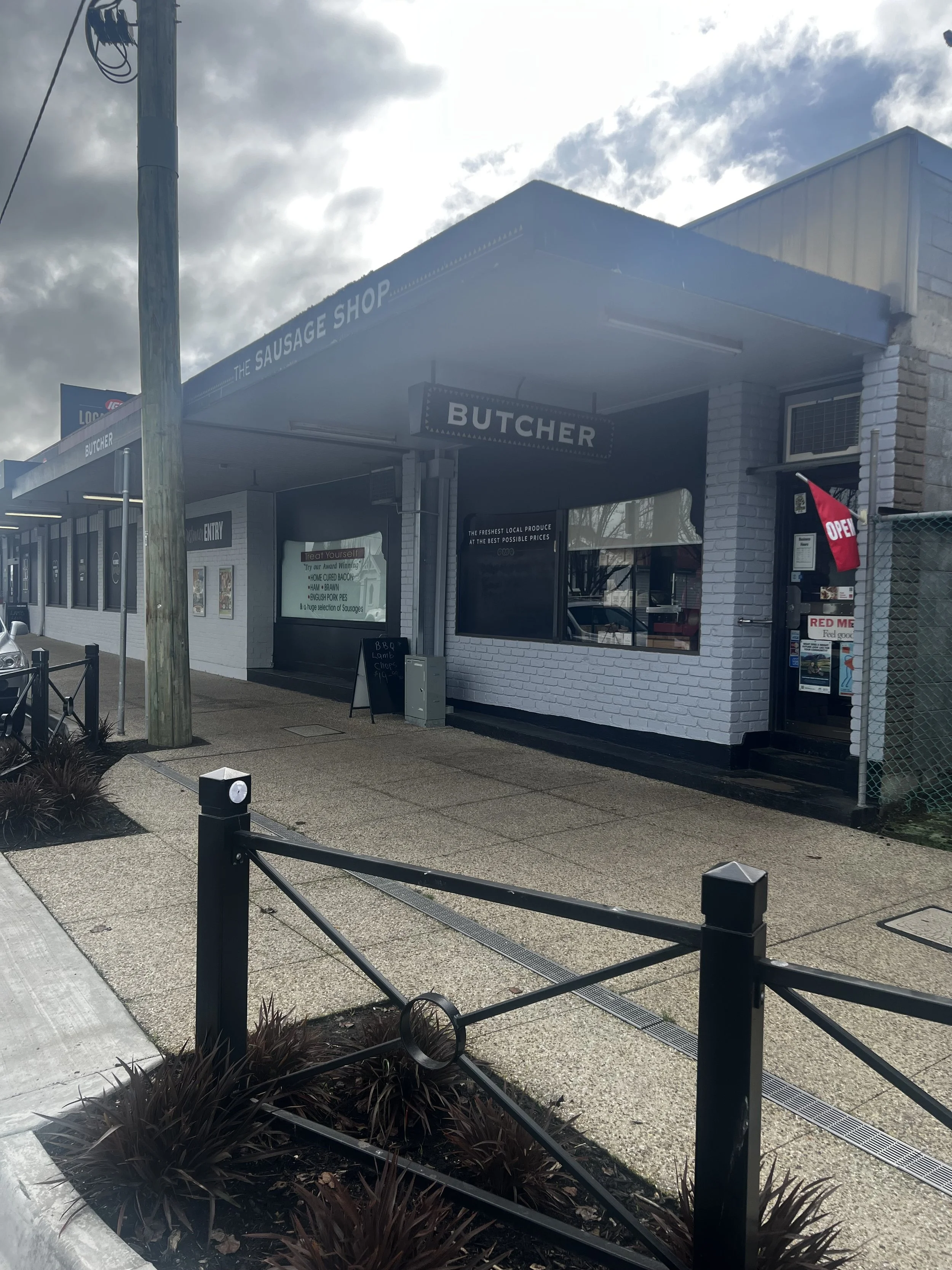 Exterior view of a butcher shop called 'The Sausage Shop' with a sign for 'Butcher' and a red 'Open' flag. There is a sidewalk with plants, black posts, and a chain in the foreground, with a cloudy sky overhead.
