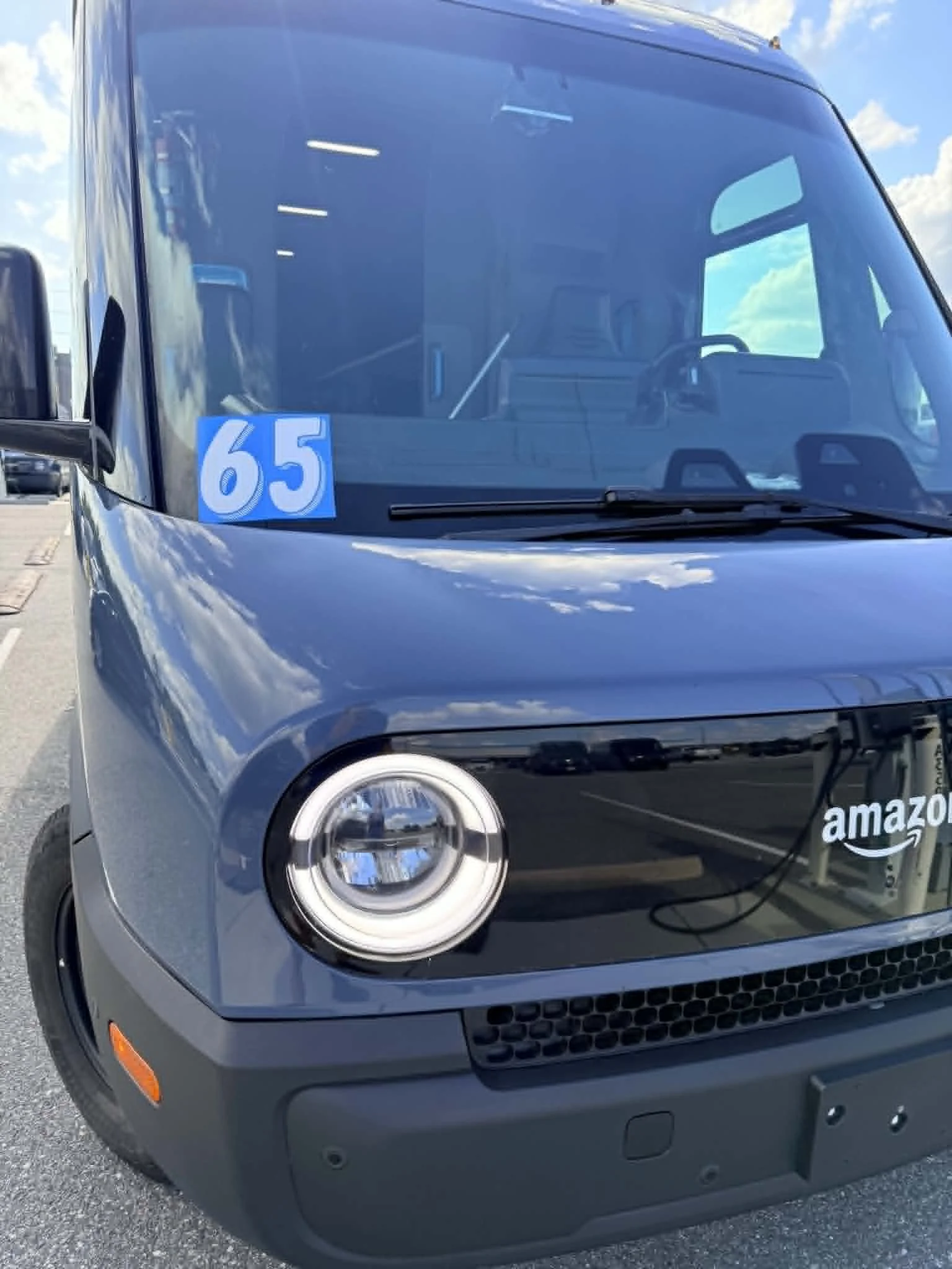 A black Amazon delivery truck parked outdoors under a blue sky with some clouds. The truck has a blue and white Amazon logo on the front. There are parking lot lines and other vehicles in the background. A blue sticker with the number 65 is on the windshield.
