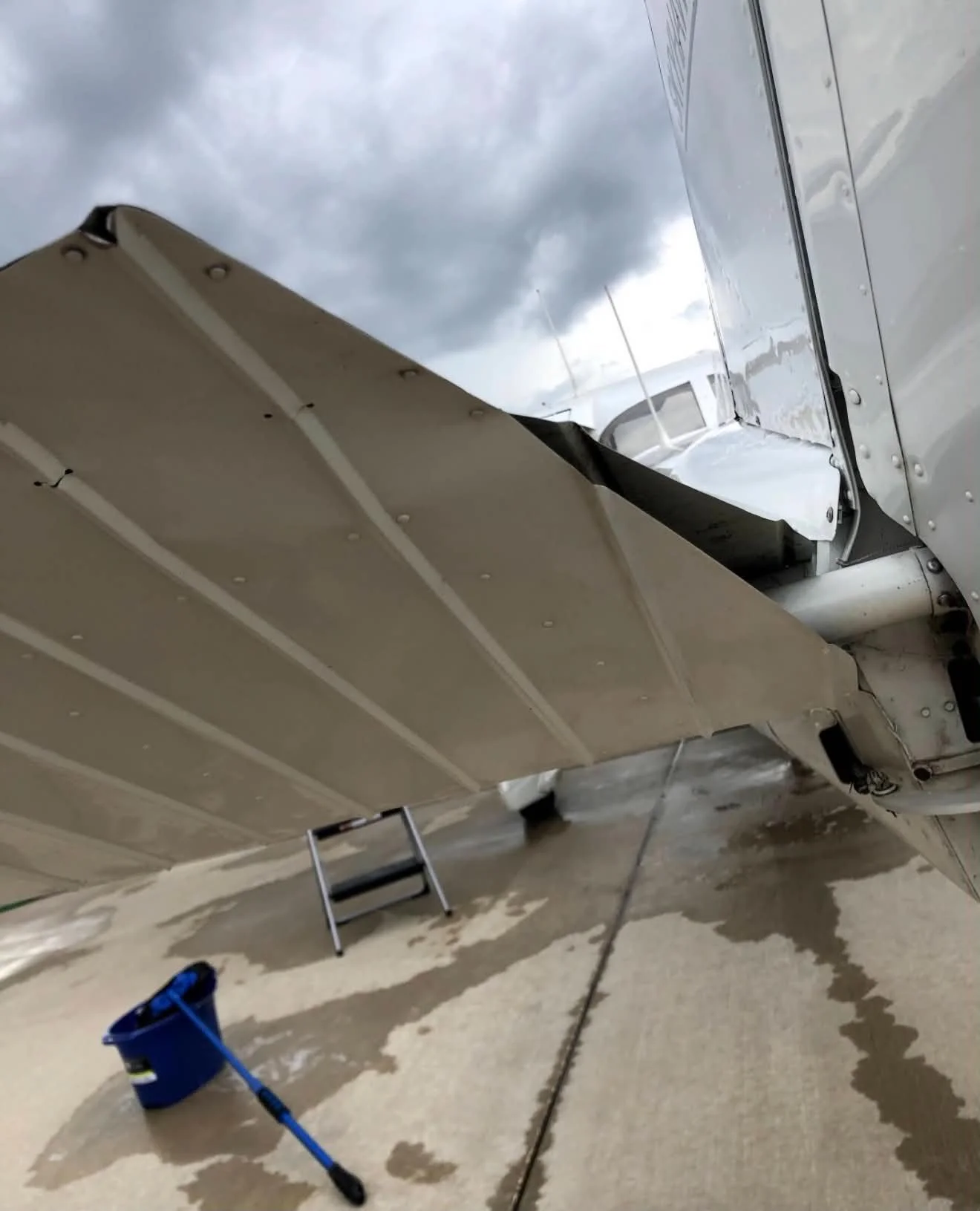 Inside view of an airplane hangar showing an aircraft wing with maintenance work, a ladder, a bucket, and water stains on the concrete floor under cloudy sky outside.