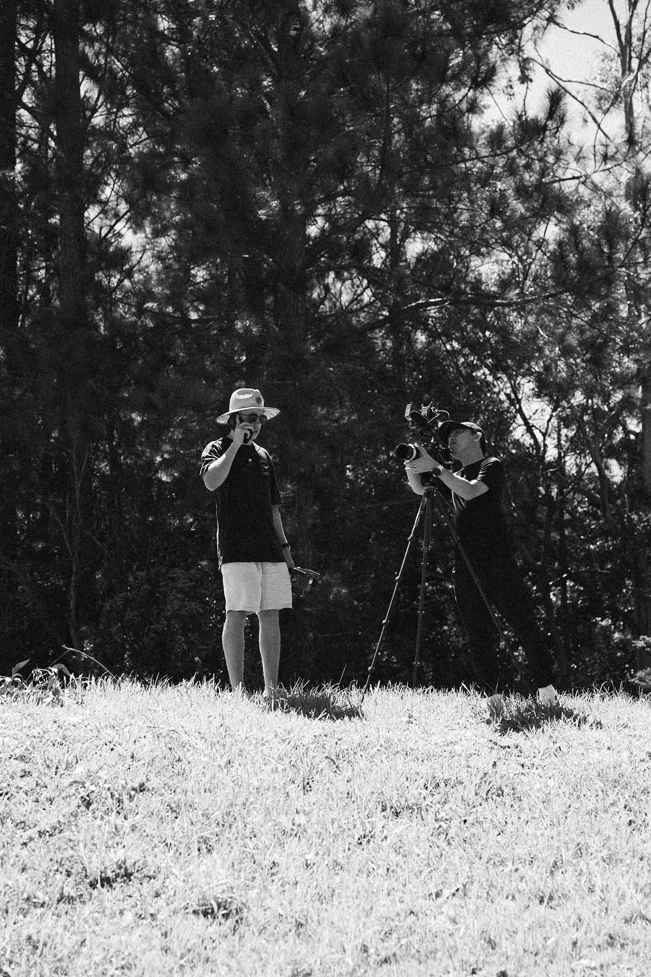 Two people, one with a wide-brimmed hat and sunglasses, and the other with a cap, are outdoors on a grassy area, one adjusting a camera on a tripod while the other drinks from a cup, with trees in the background.
