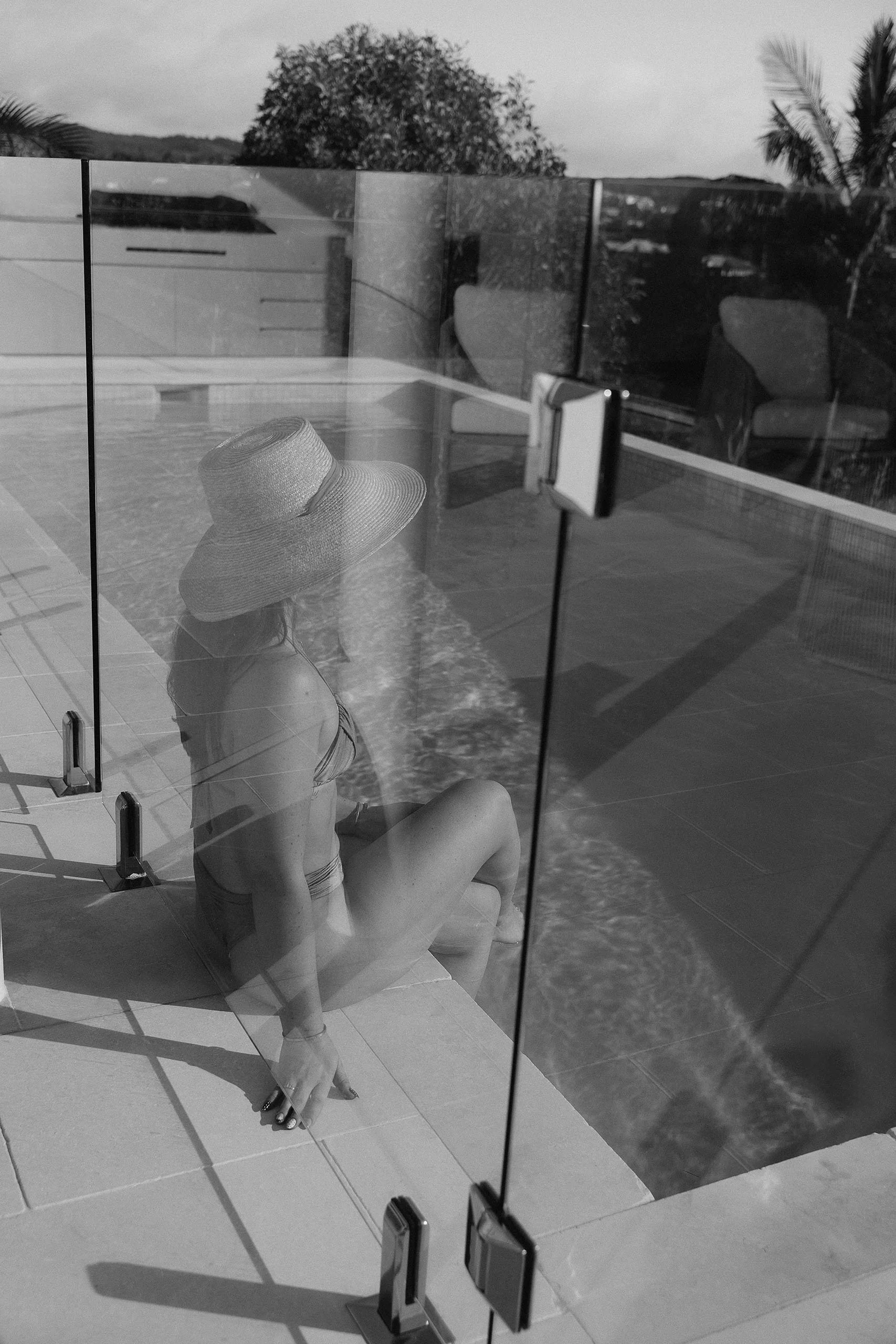 Woman in a sunhat seated beside the pool behind frameless glass fencing at Horizon House, Sunrise Beach.