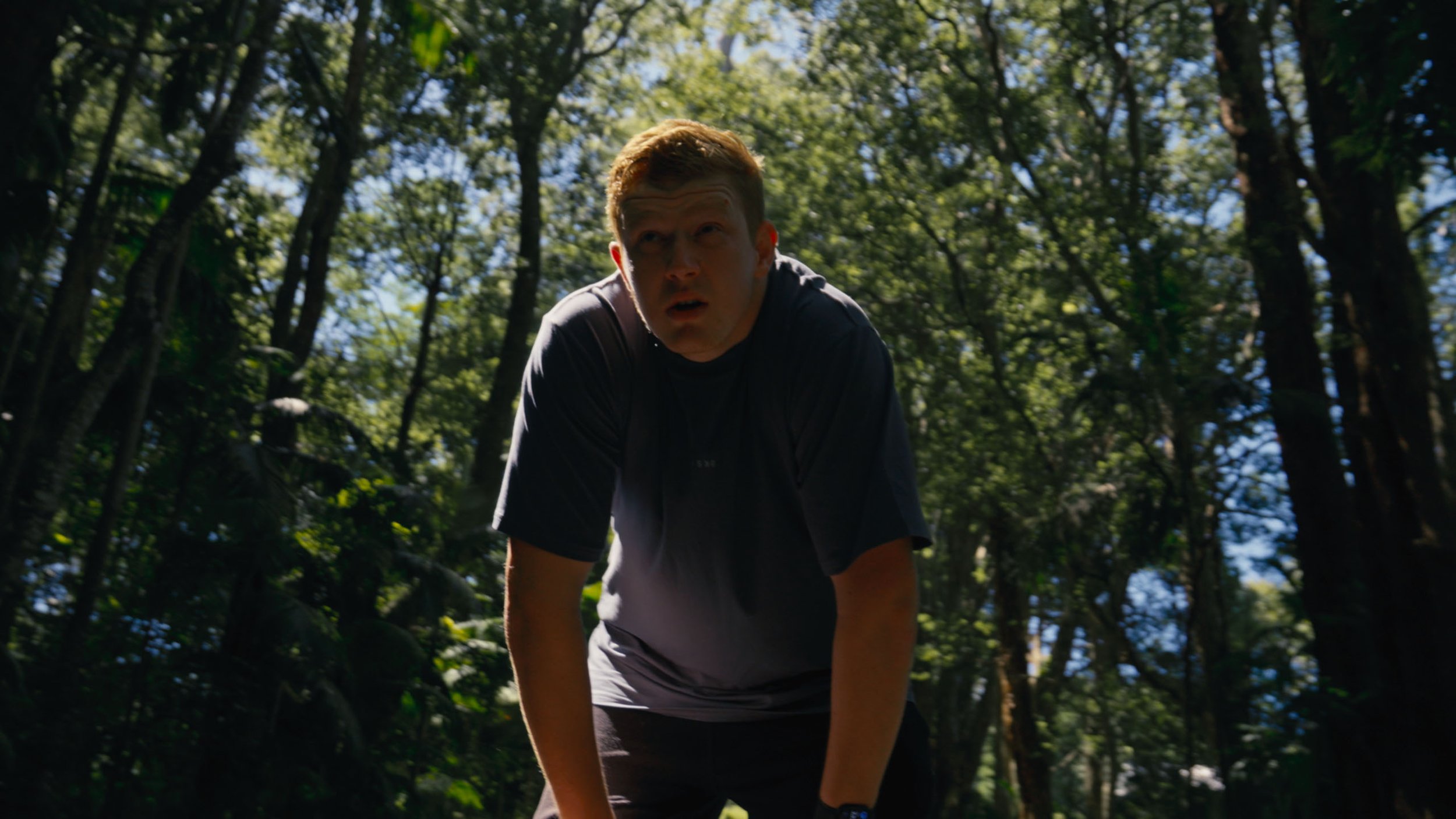 A young man with short hair, wearing a black shirt, leaning forward outdoors in a wooded area with tall trees and sunlight filtering through the leaves.