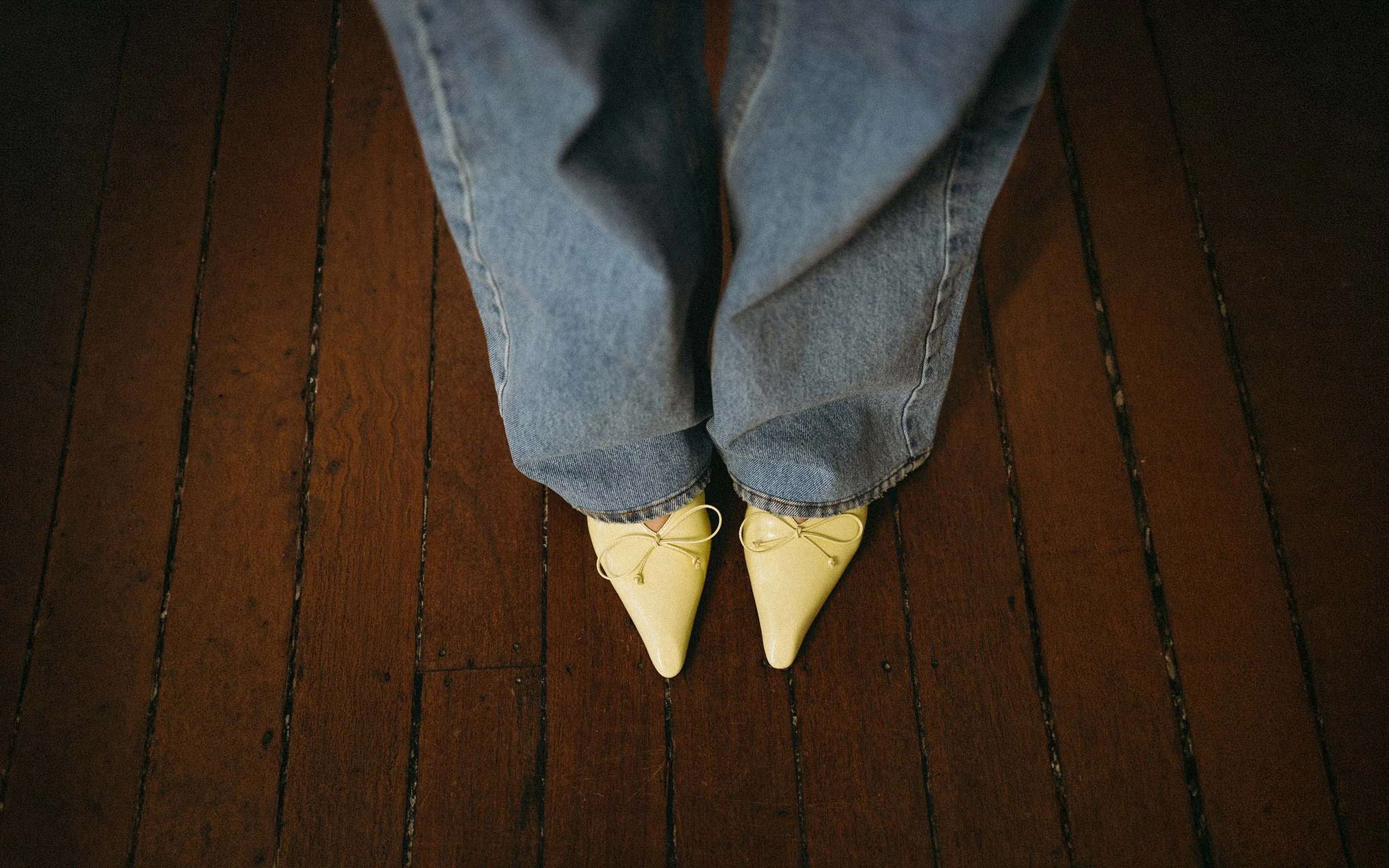 Person wearing light yellow pointed shoes, blue jeans, standing on a dark brown wooden floor.