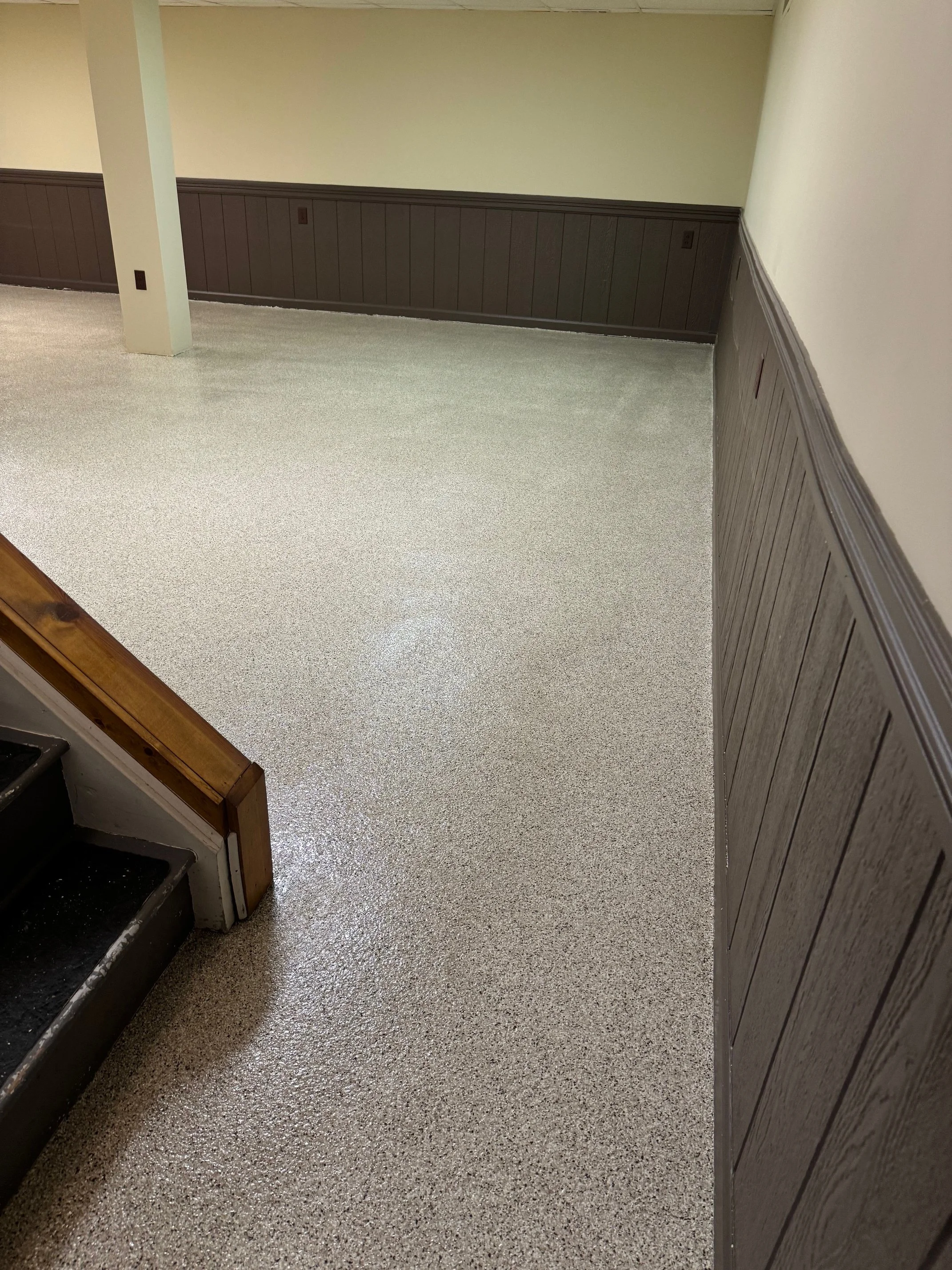 Basement with brown paneling on the walls, a stairway to the left, and a grey speckled epoxy floor