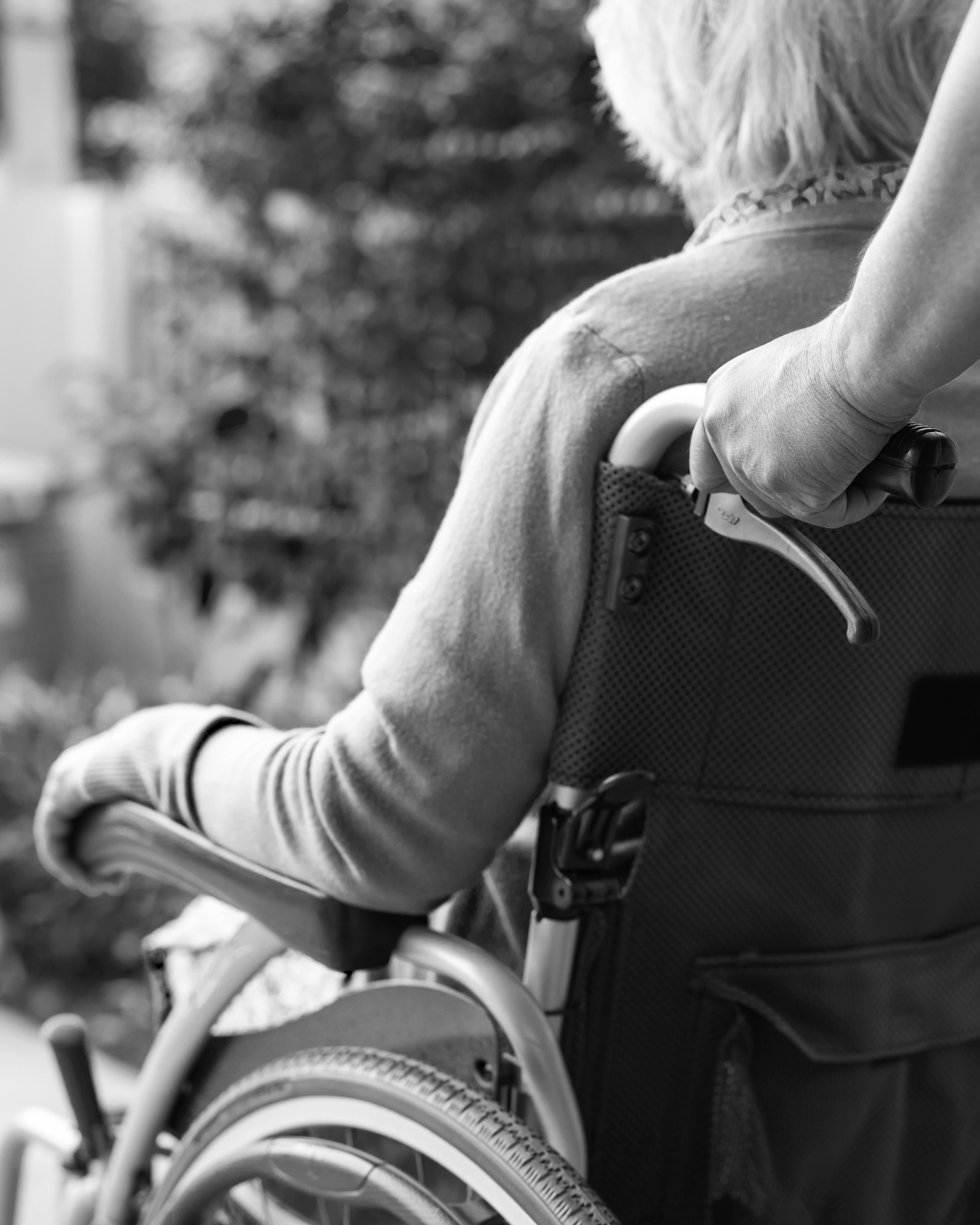 Close-up of a person in a wheelchair holding an IV pole outside near trees, in black and white.