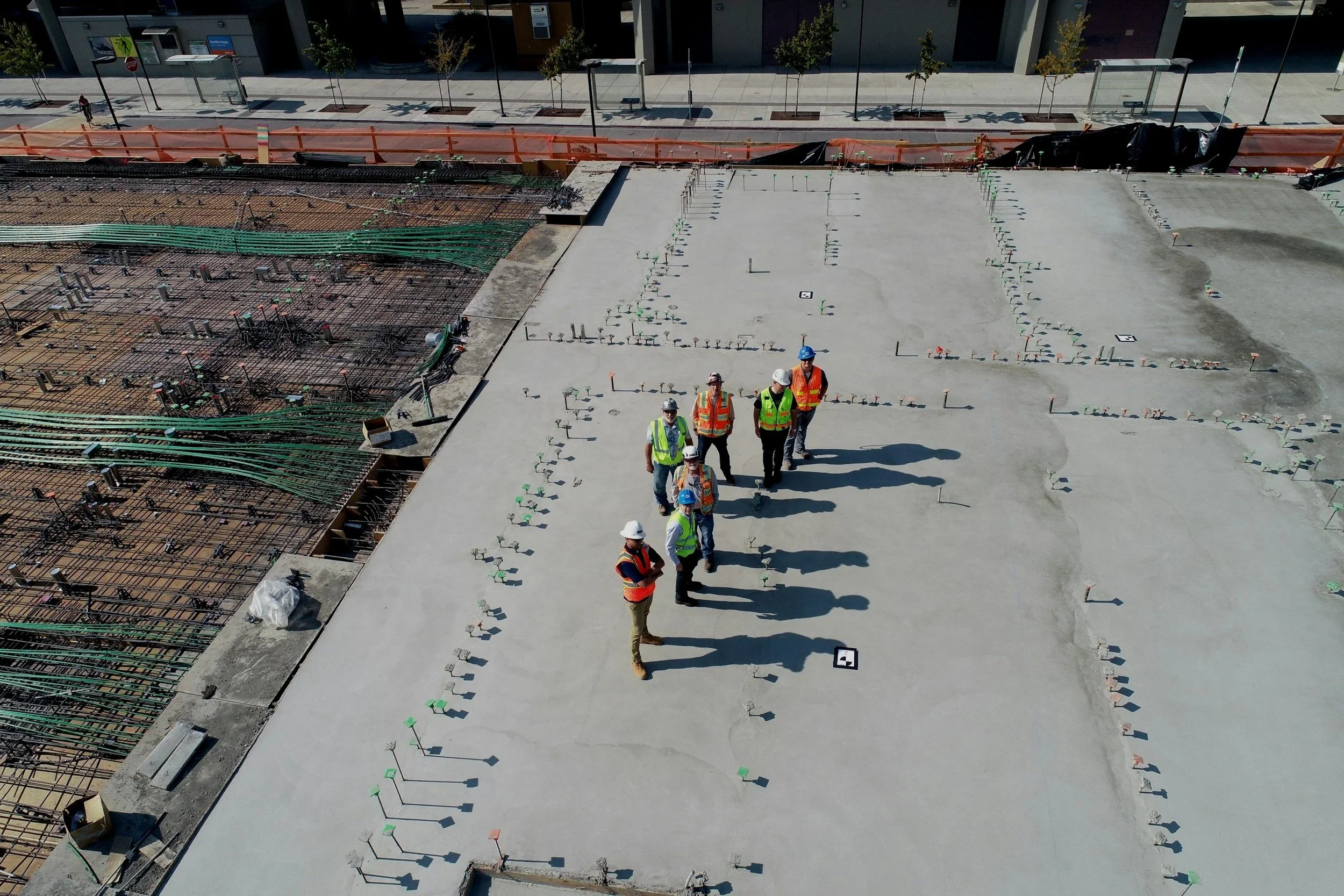 Construction site with workers in safety vests and helmets walking on a concrete slab. Reinforcement bars are visible on the left side, and various small equipment and markings are seen on the ground. A city sidewalk with trees and buildings is in the background.