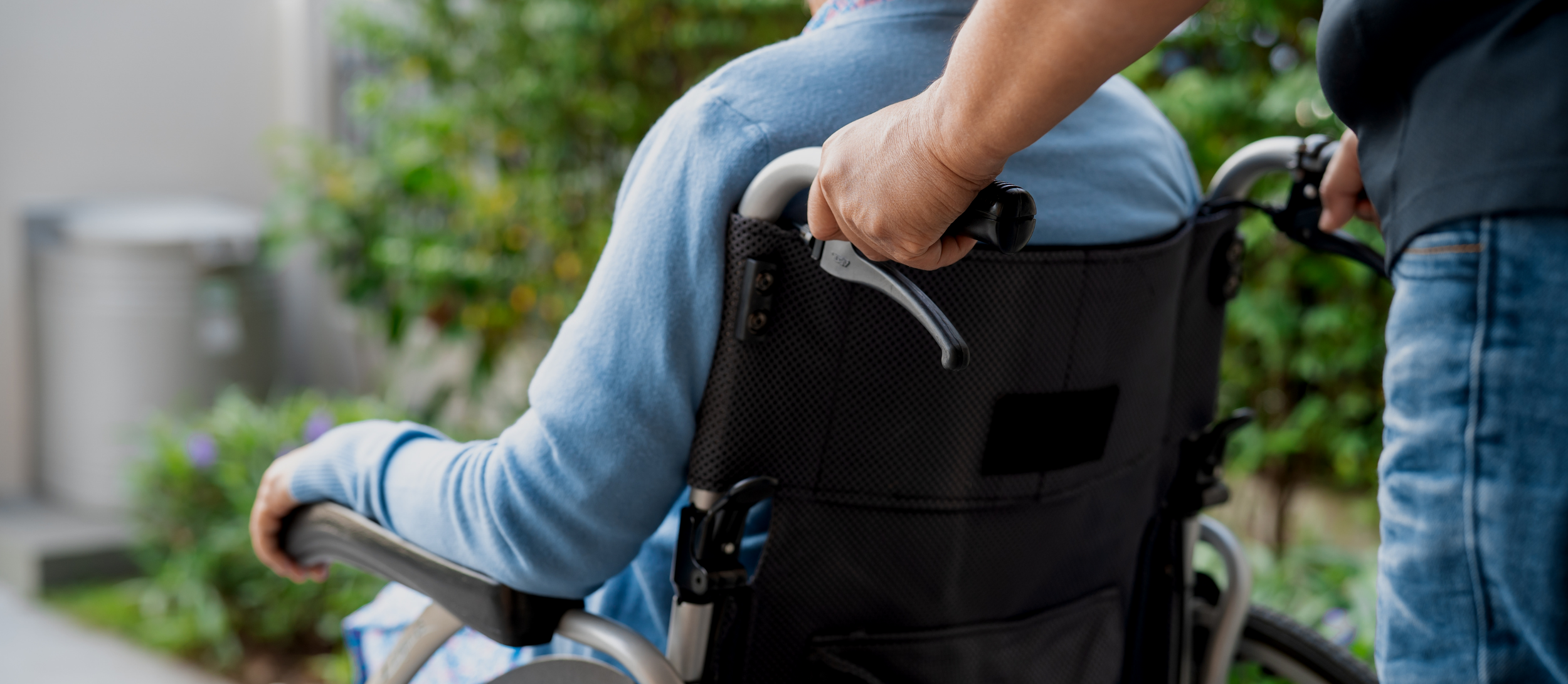 Close-up of a person in a blue long-sleeve shirt sitting in a wheelchair, with another person holding the wheelchair handle outdoors.
