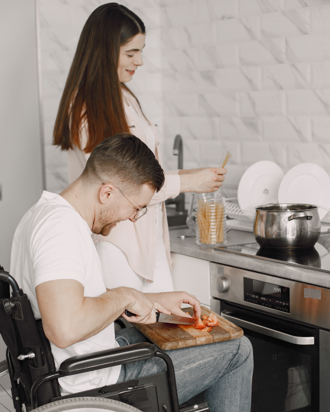 A man in a wheelchair is chopping tomatoes on a wooden cutting board in a kitchen. A woman in the background is preparing food with spaghetti in a jar.