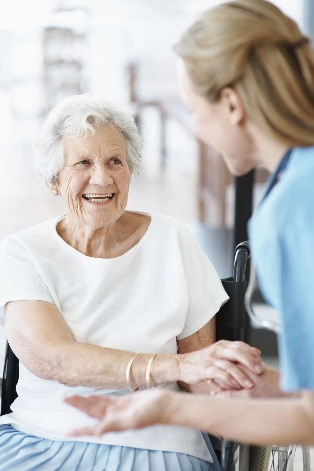An elderly woman with white hair smiling and holding hands with a caregiver in a healthcare setting.